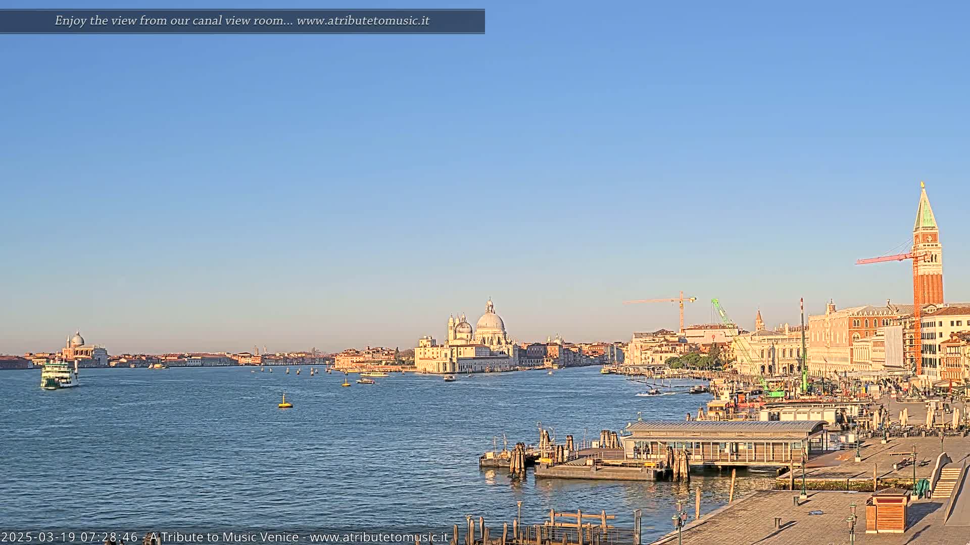 A calm, clear day reveals a Venetian waterway with boats, a distant church, and buildings lining the shore, under a pale blue sky.
