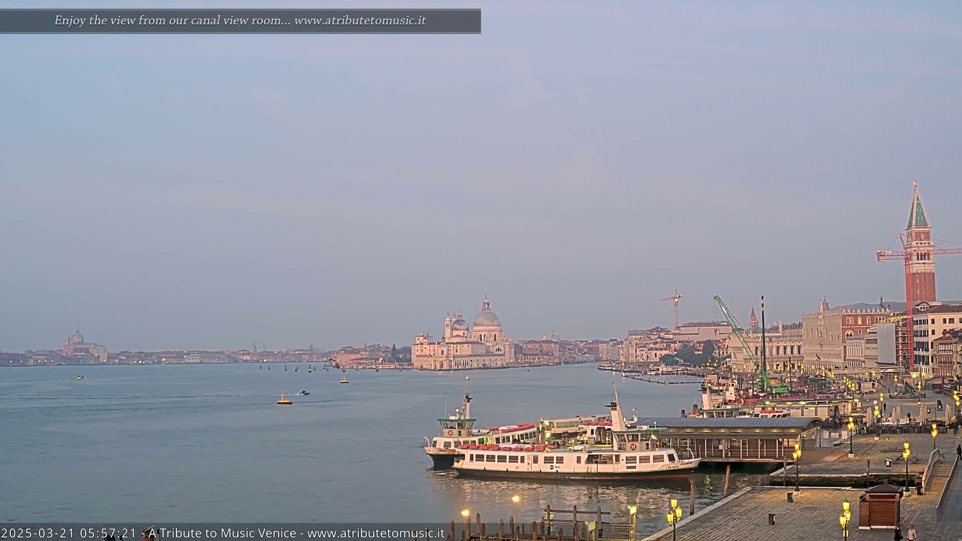 A calm, pale-blue dawn illuminates a Venetian canal scene featuring a ferry docked near a waterfront promenade, with the Basilica di Santa Maria della Salute and St. Mark's Campanile visible in the distance.