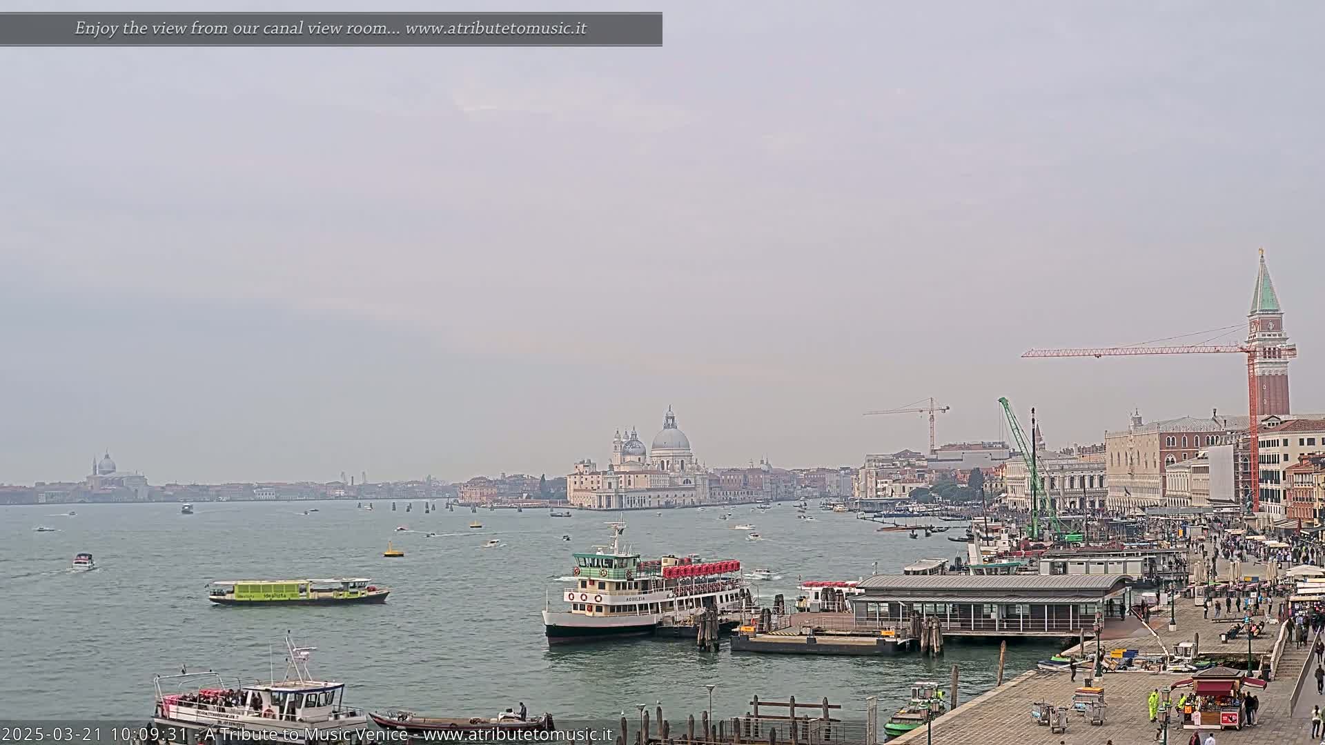 A wide shot of a Venetian canal shows several boats, a waterfront building, and the city skyline under an overcast sky.