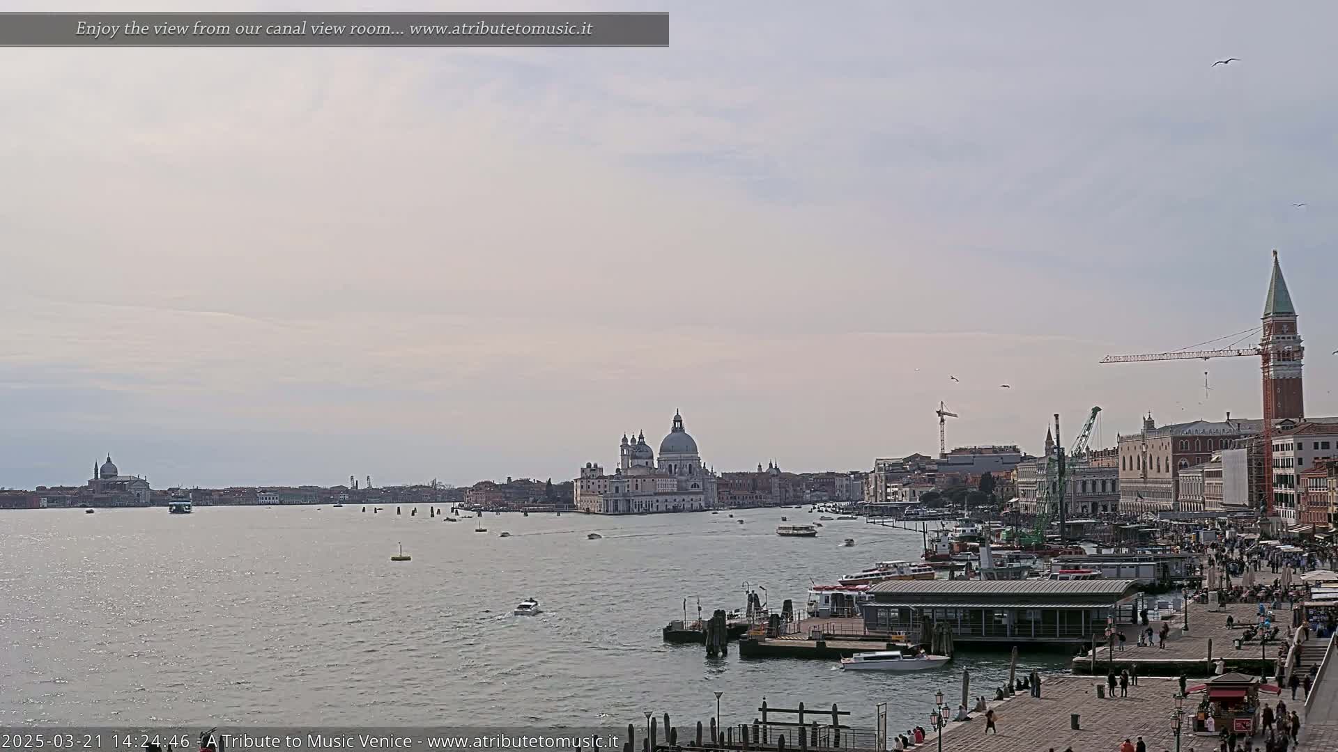 A wide, calm canal scene in Venice under a pale, overcast sky shows a cityscape with boats, a large domed church, and a tall red brick tower.