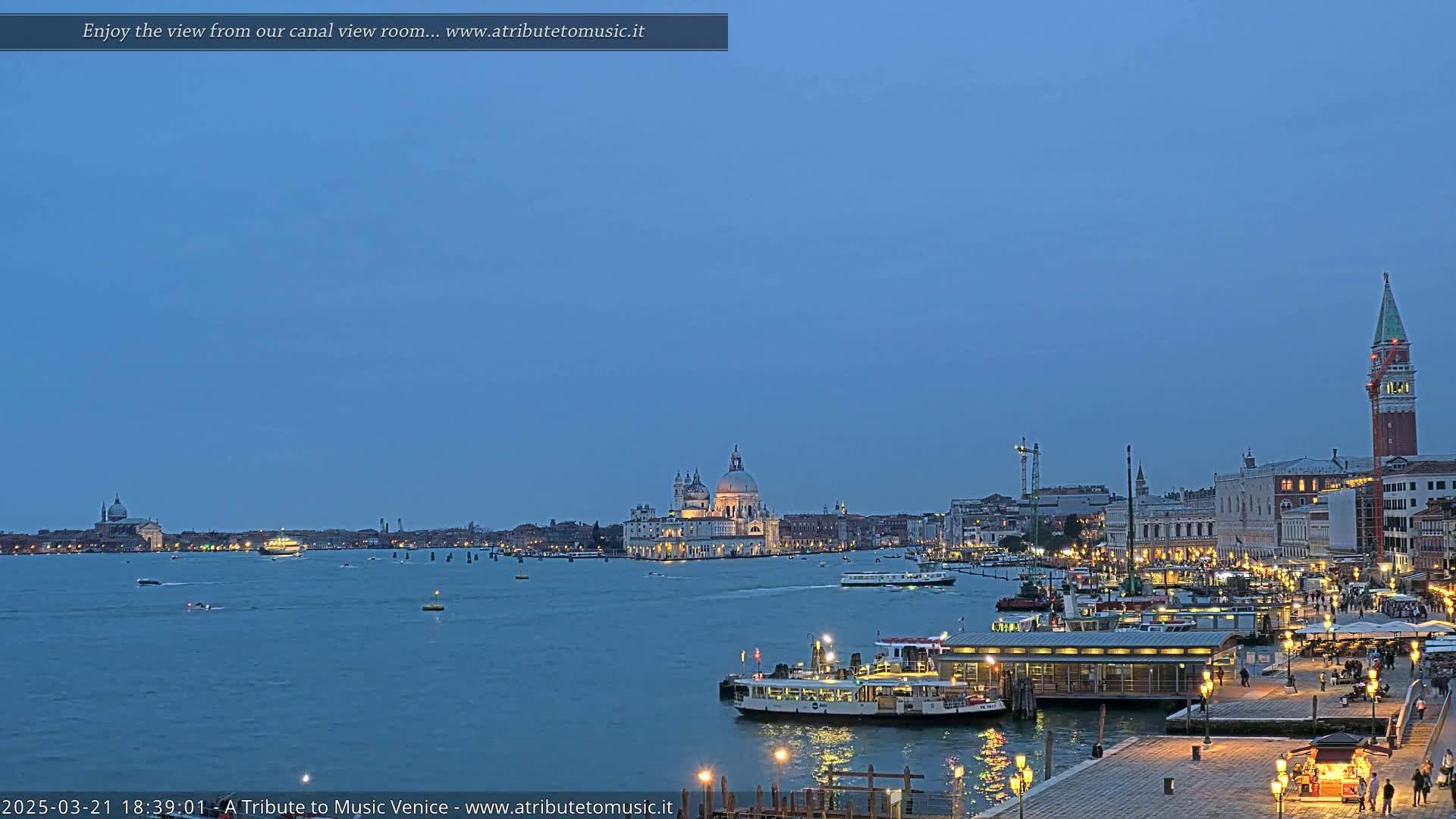 A twilight view of a canal in Venice, showing illuminated buildings, boats, and a large church in the distance under a clear, dark-blue sky.