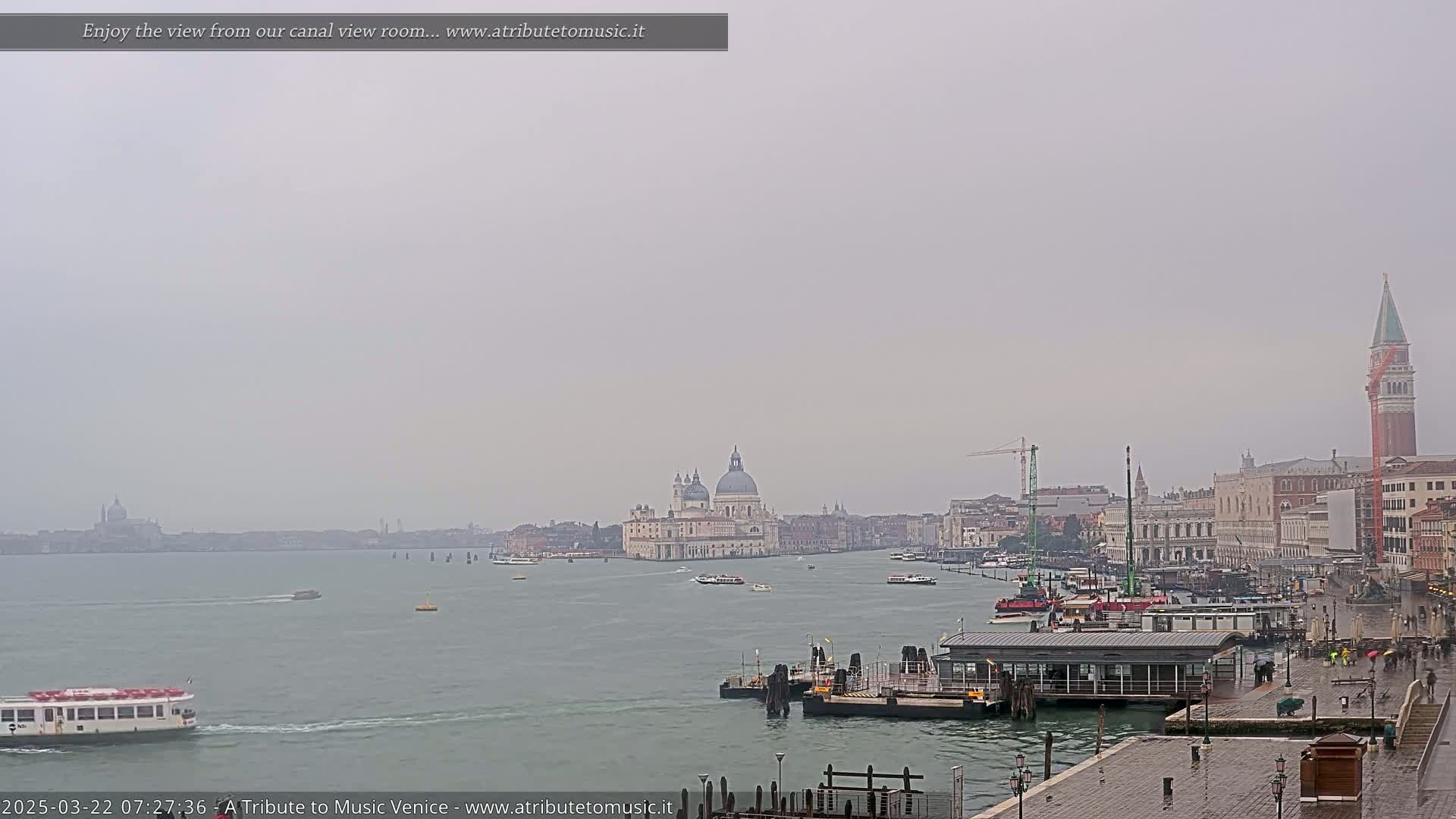 A hazy, rainy view of Venice shows a waterway with boats, a distant basilica, and buildings along the canal.