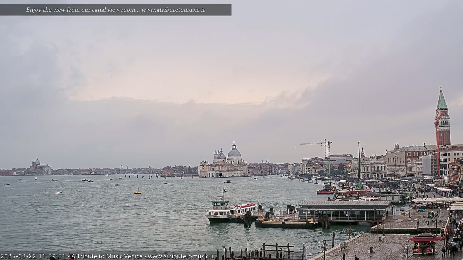 A wide shot of a waterway in Venice, Italy, on an overcast day shows a ferry and other boats, along with the city skyline including the Basilica di Santa Maria della Salute and St. Mark’s Campanile.