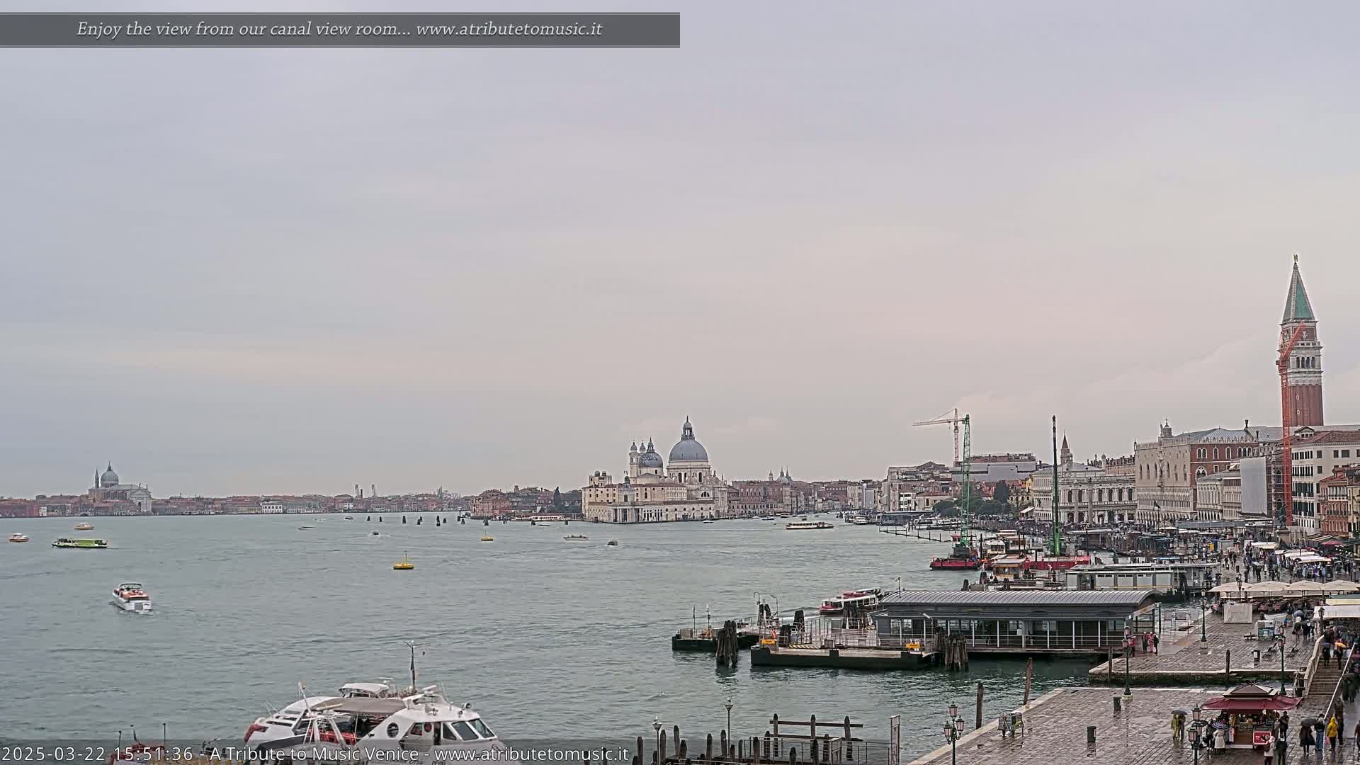 A wide, calm body of water shows numerous boats and a distant cityscape under an overcast sky, featuring a large domed church and a tall bell tower.