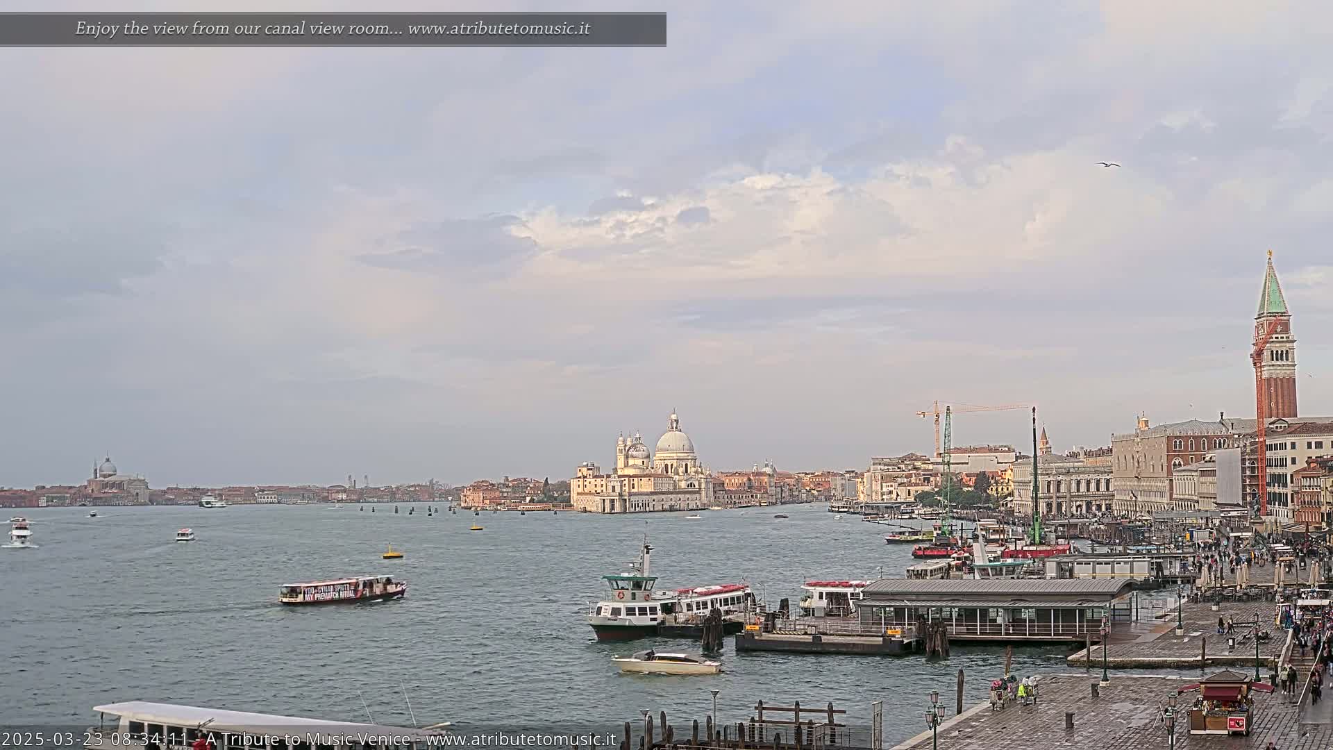 A wide shot of a Venetian canal shows several boats and buildings under a mostly cloudy sky.
