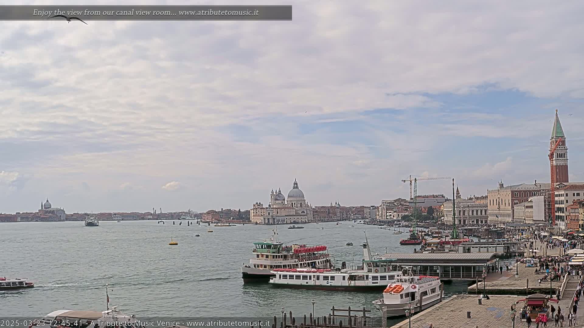 A partly cloudy sky overlooks a Venetian canal with several water taxis and a cityscape featuring a large dome and bell tower.