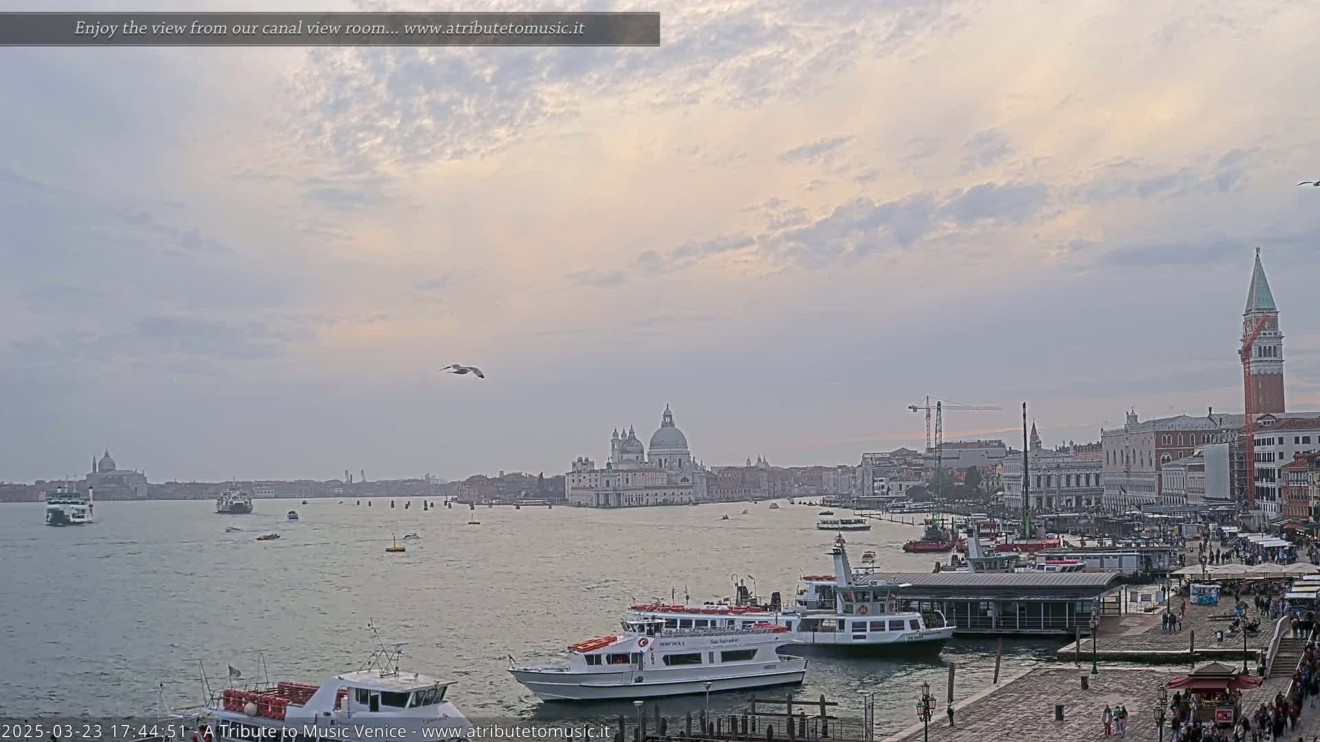 A wide shot of a Venetian canal shows several boats, a cityscape including the Basilica di Santa Maria della Salute, and a cloudy, light-pink sunset sky.