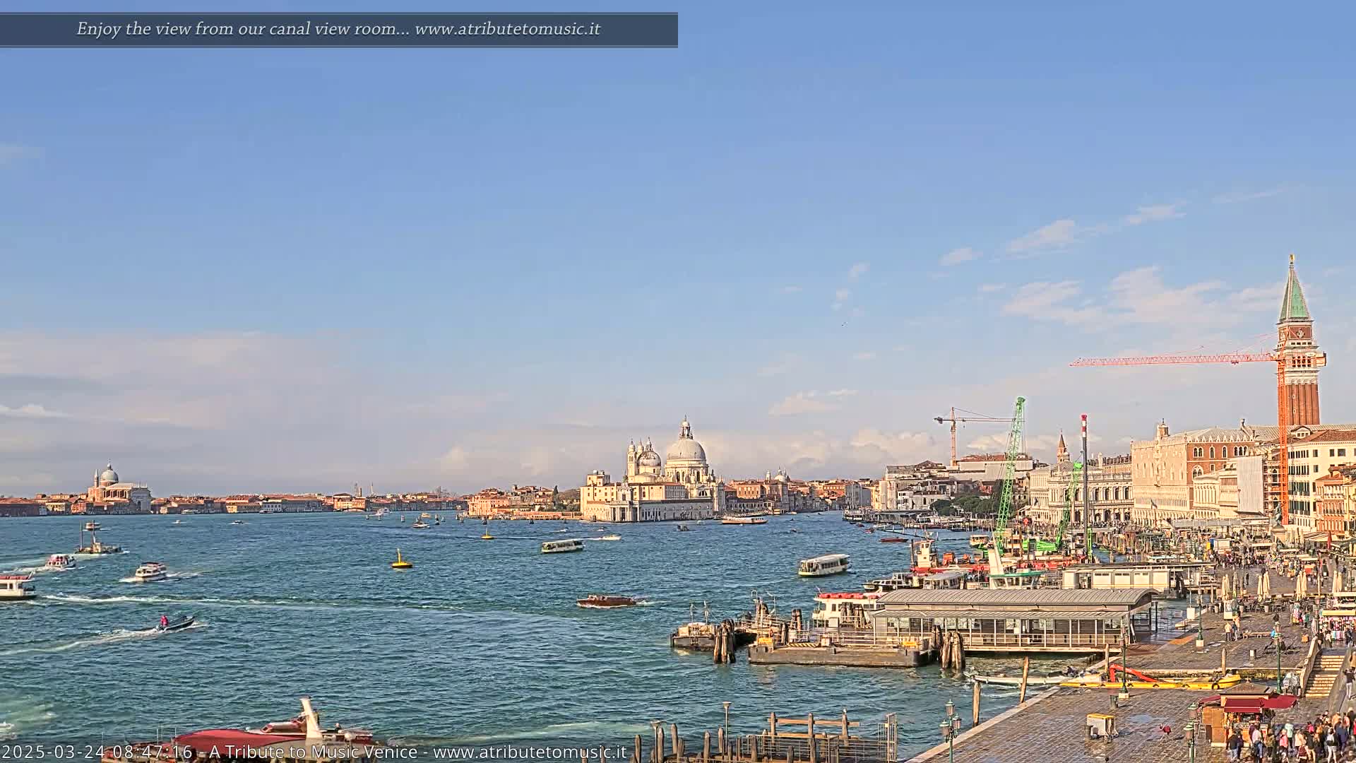 A wide shot of a Venetian canal on a sunny day shows numerous boats, a large church in the distance, and a city waterfront with people and construction cranes.