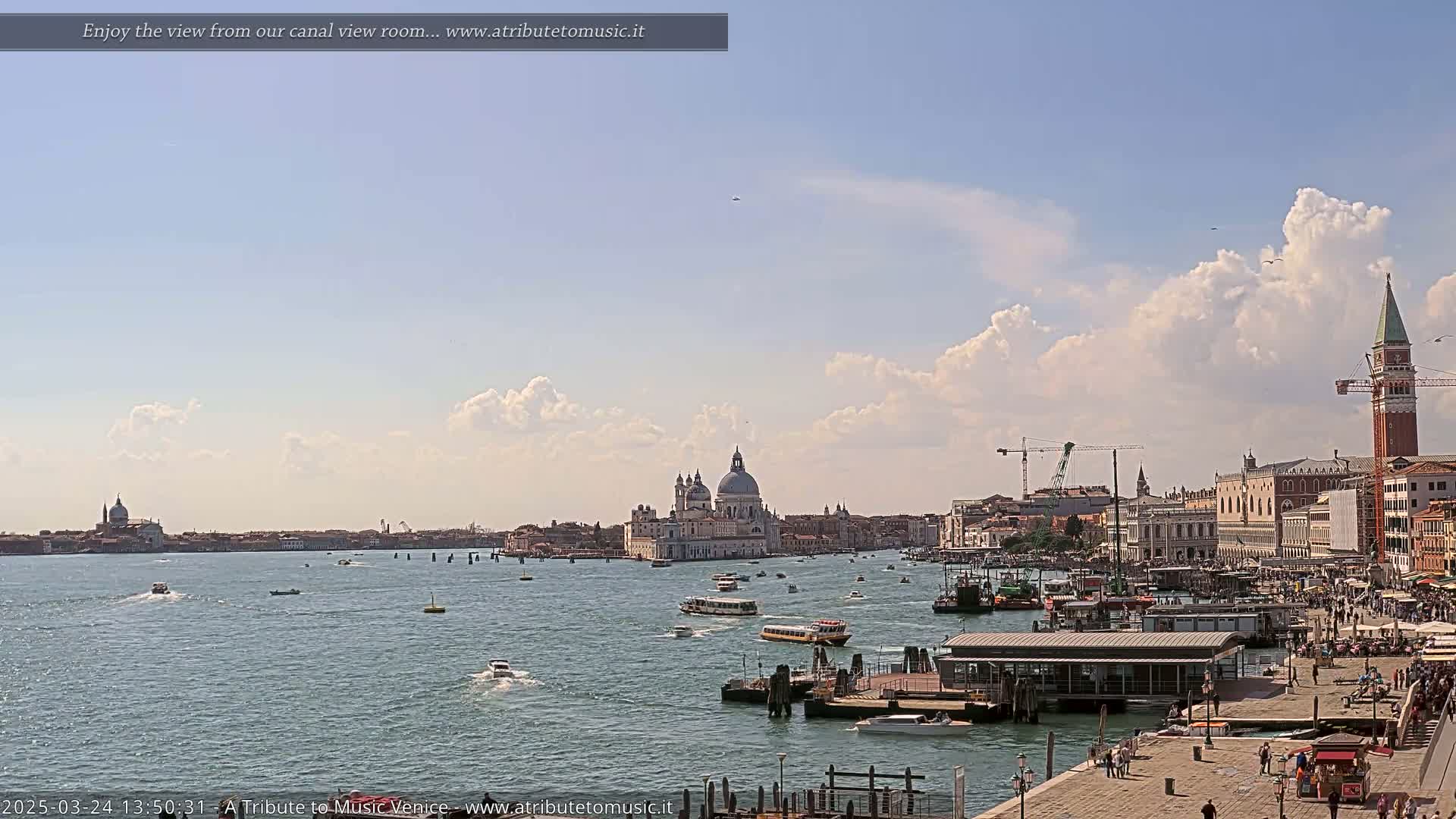A wide shot of a Venetian canal on a partly sunny day with fluffy white clouds shows boats on the water, buildings lining the canal, and a large church in the distance.