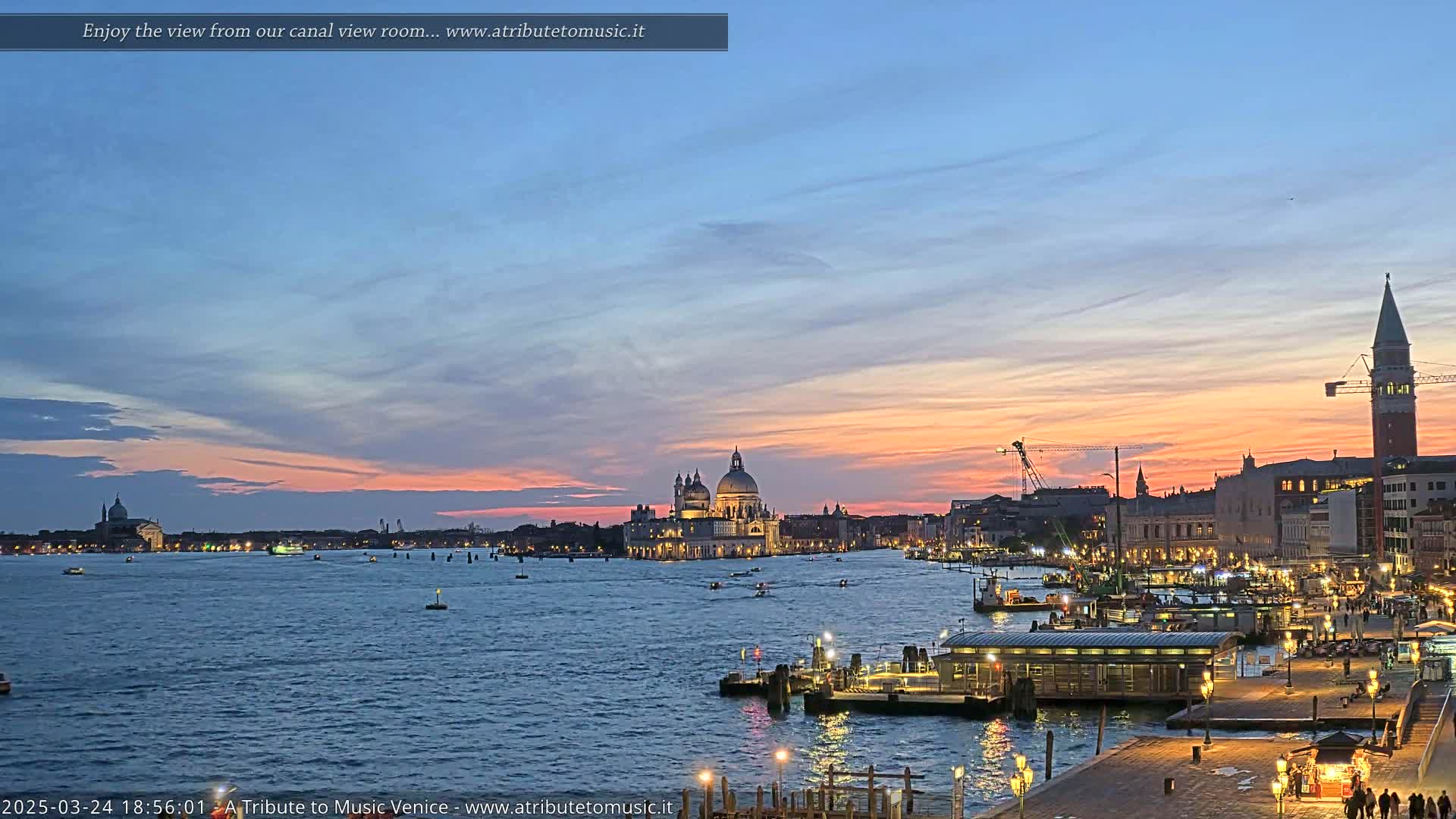 A Venetian sunset over a calm canal, with illuminated buildings and boats, features a large domed church and a tall bell tower in the distance.