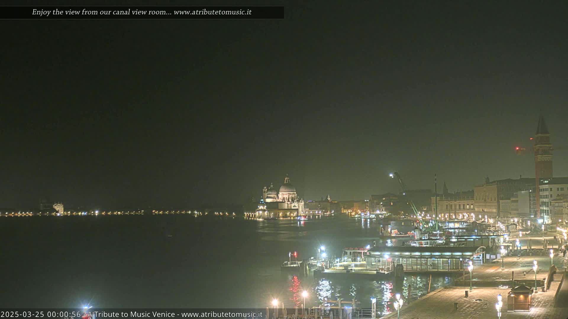 A nighttime view of a canal in Venice, showing illuminated buildings and boats under a dark, hazy sky.