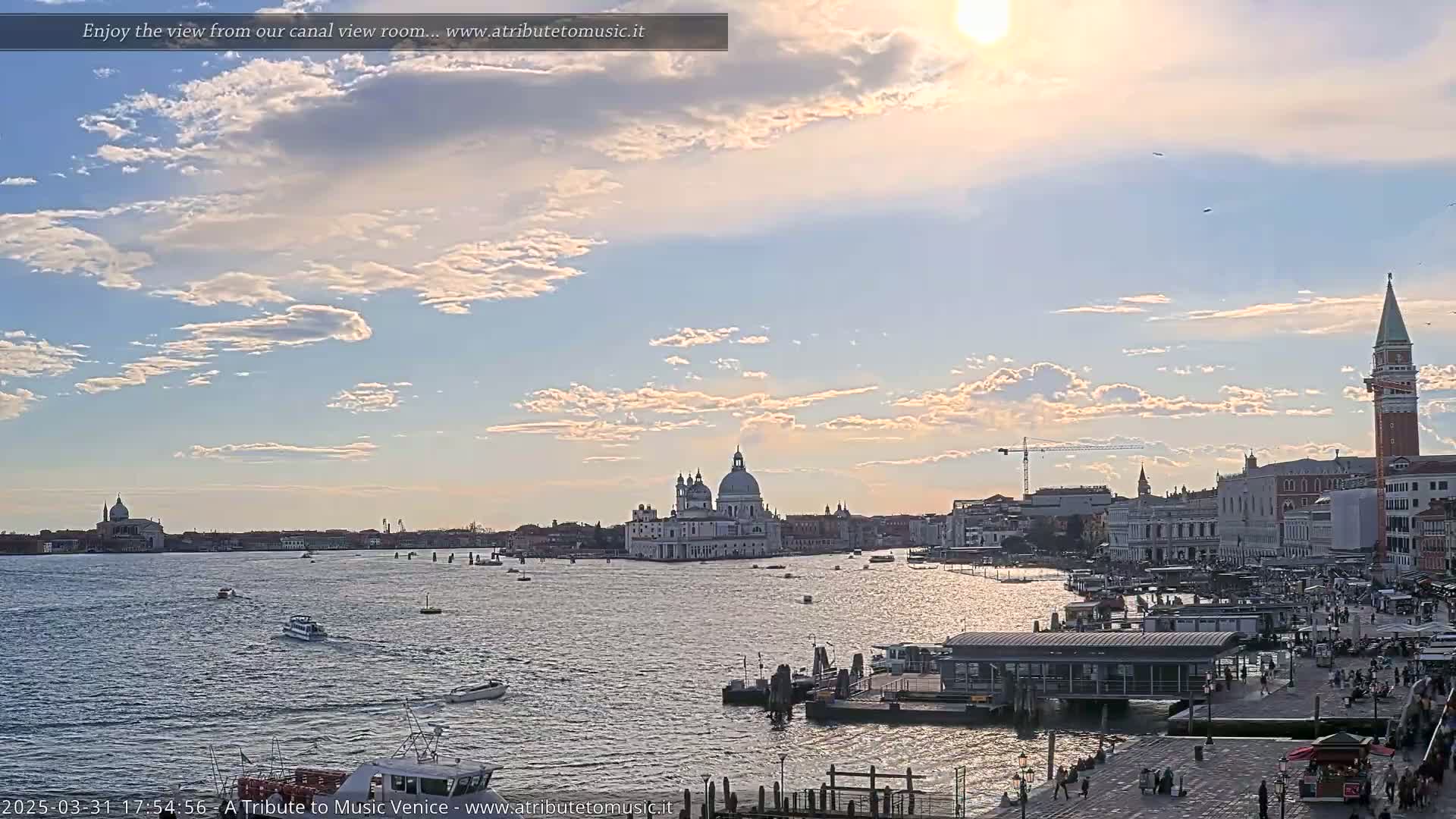 A sunny day with scattered clouds overlooks a Venetian canal, showing a cityscape with a large white domed church and a tall red brick tower.