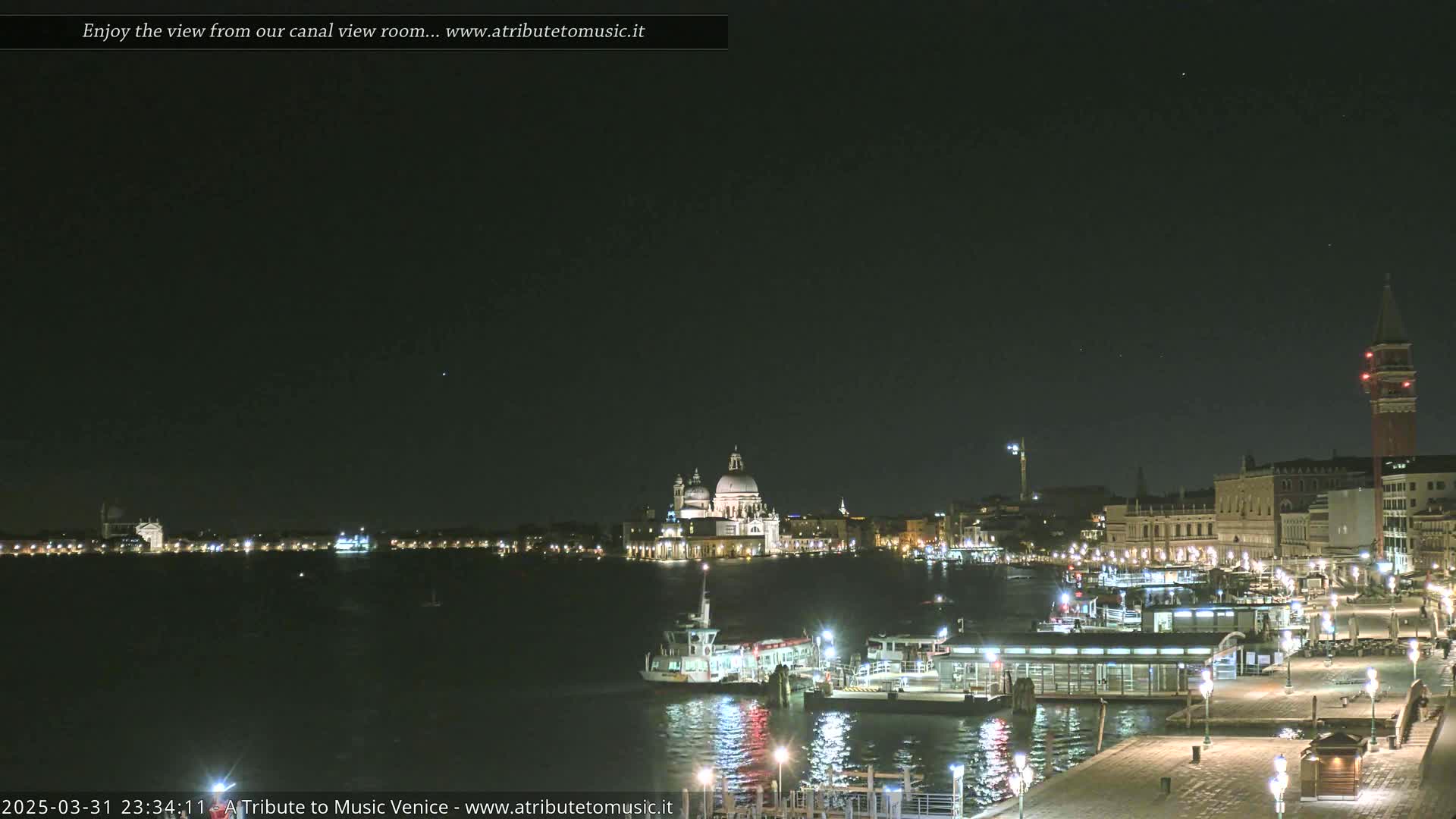 A nighttime view of a canal in Venice, Italy, shows buildings and boats illuminated along the water's edge under a clear, dark sky.