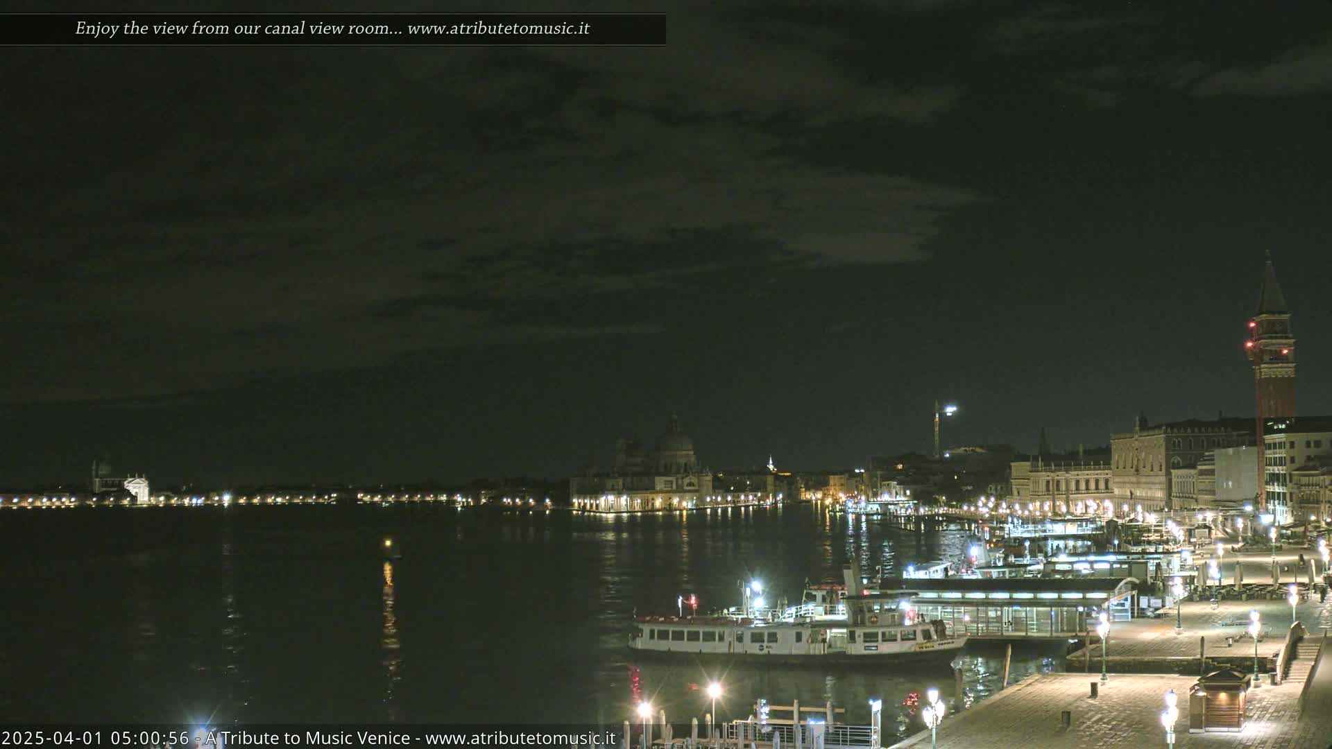 A nighttime view of a canal in Venice, showing illuminated buildings and boats under a partly cloudy sky.