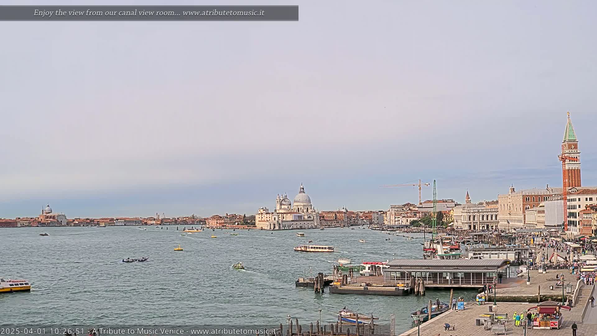 A wide shot of a Venetian canal, under a mostly cloudy sky, shows numerous boats, a waterfront building, and the distant Basilica di Santa Maria della Salute.