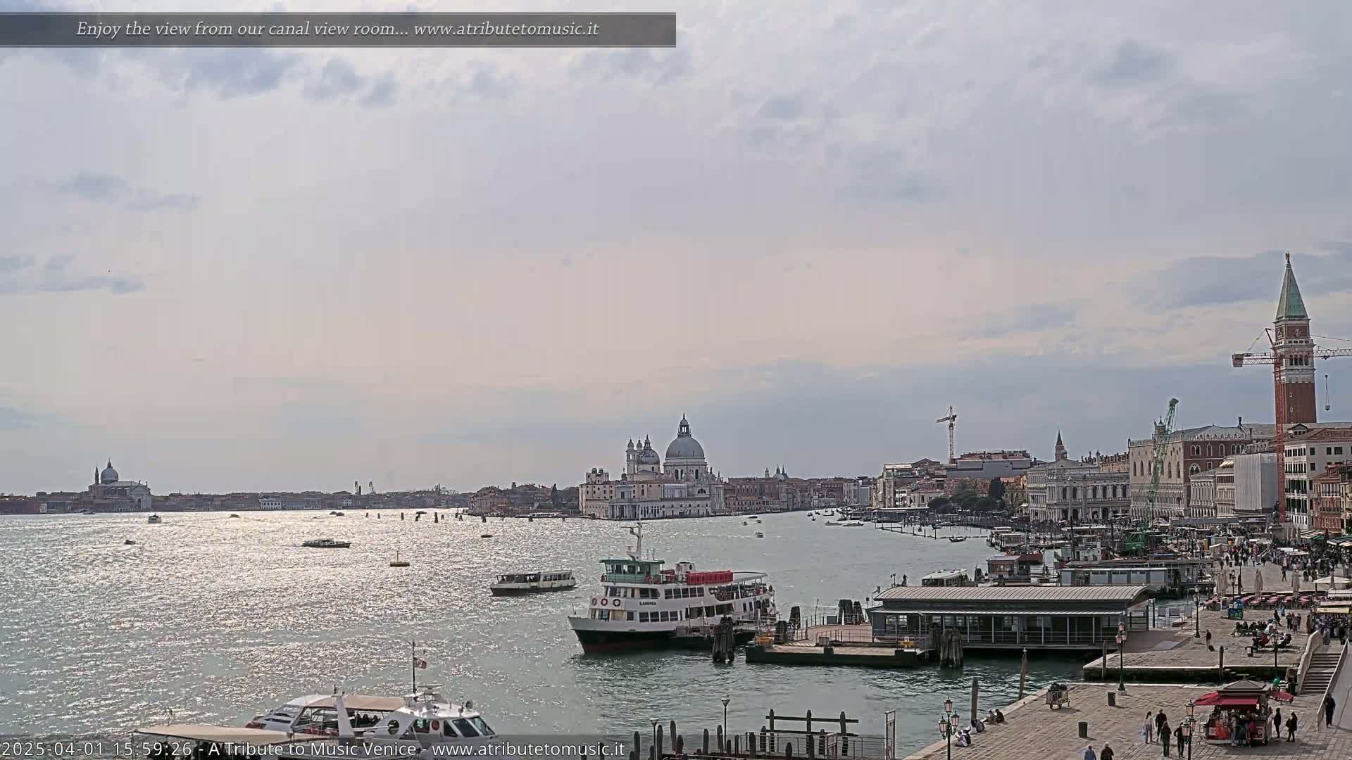 A sunny day in Venice shows a canal with several boats, including a large passenger ferry, and a cityscape featuring prominent buildings and a tall bell tower.
