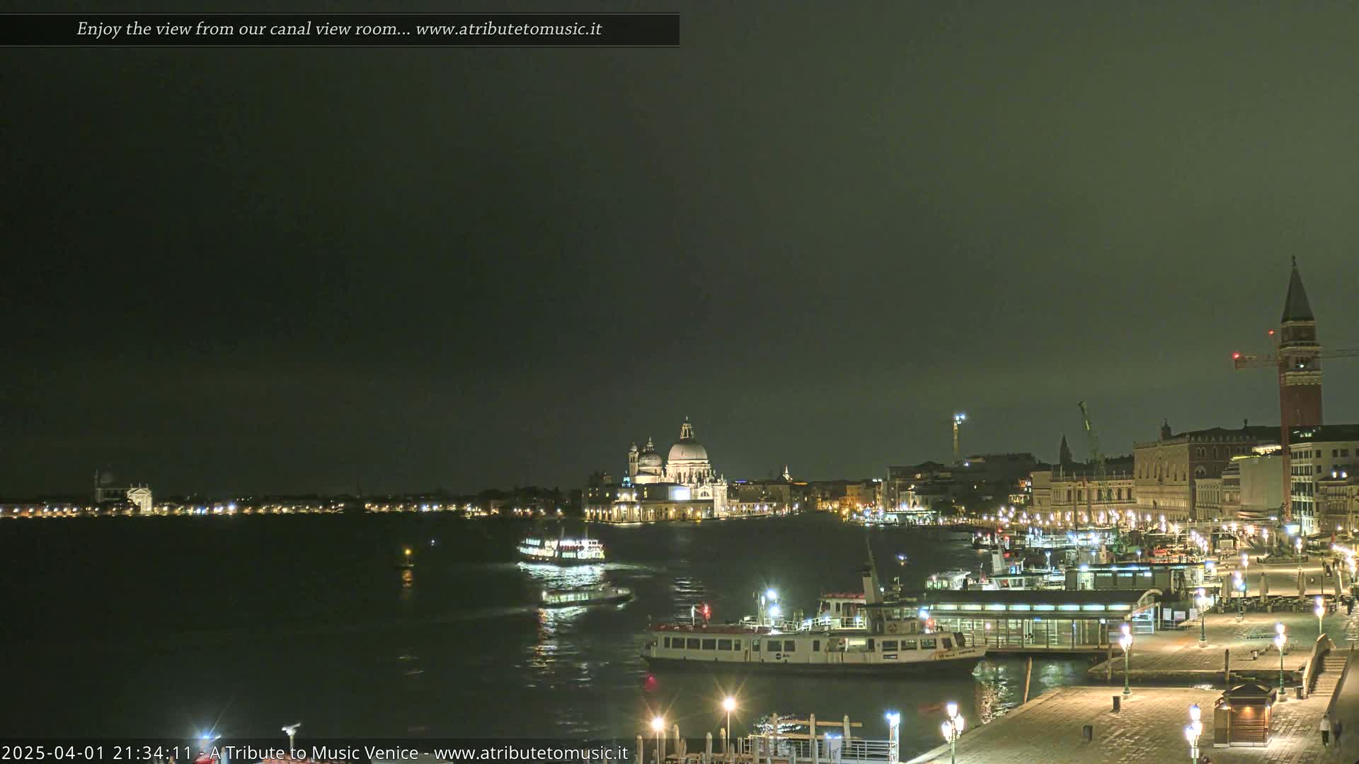 A nighttime view of a canal in Venice, Italy, shows several boats docked near a waterfront promenade with buildings and a large church in the background under a dark, cloudy sky.