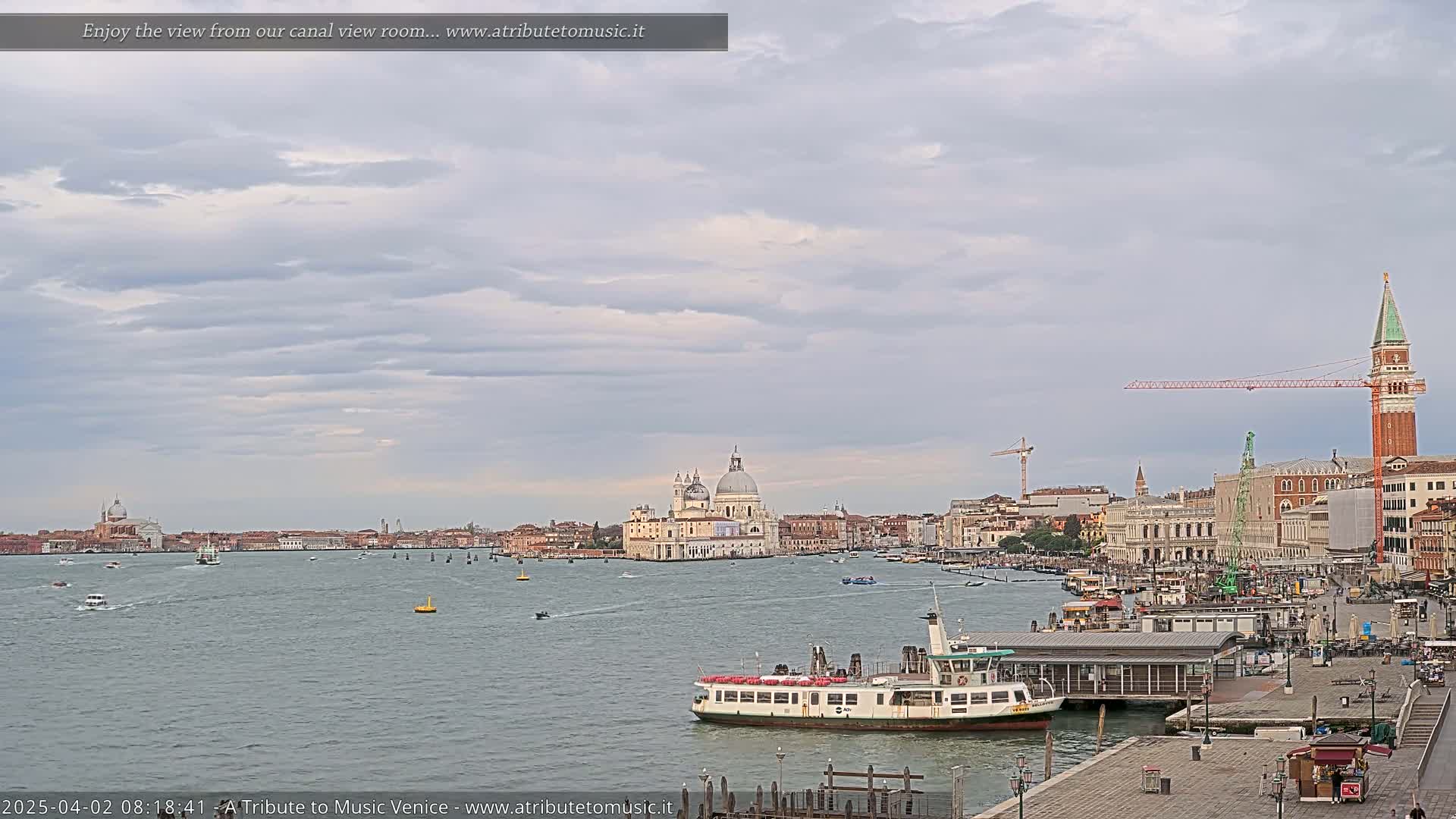 A ferry is docked near a waterfront in Venice, Italy, under a cloudy sky, with the Basilica di Santa Maria della Salute visible across a calm body of water.