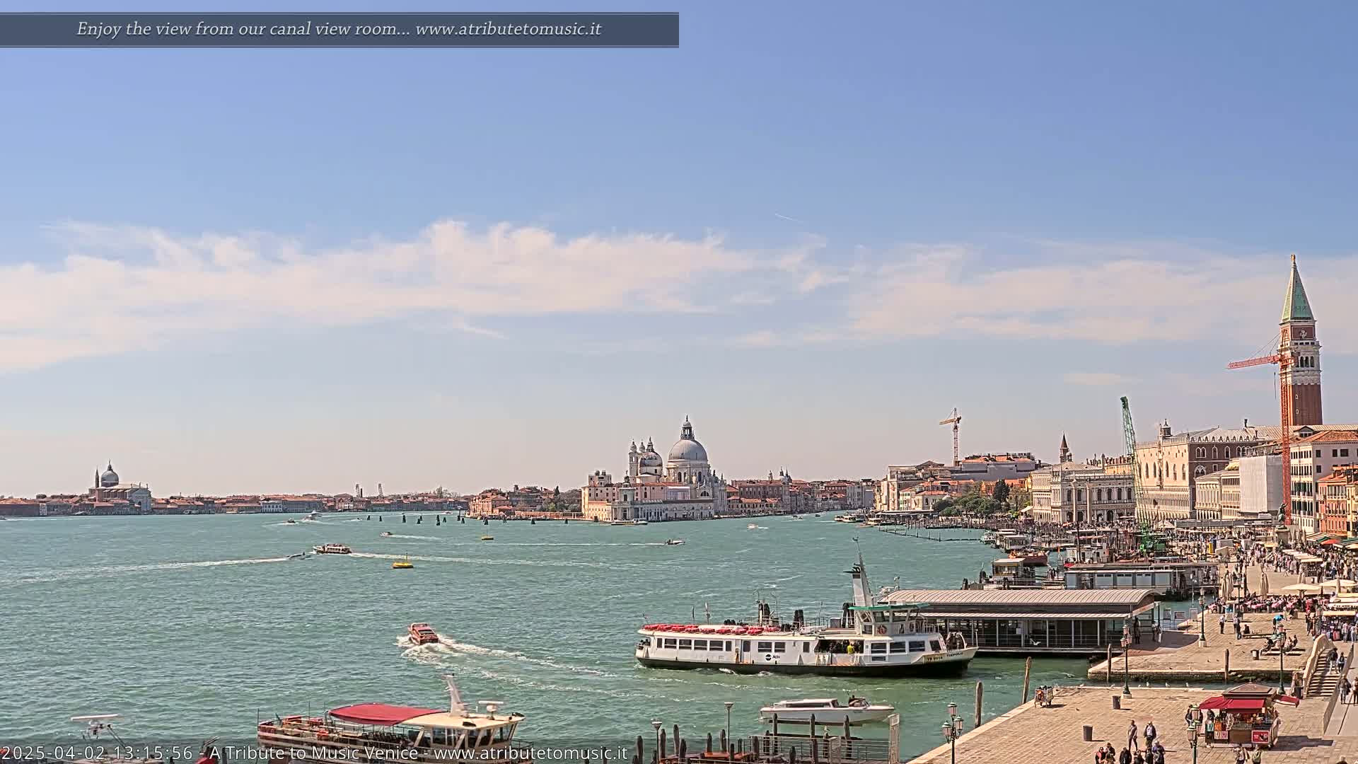 A sunny day in Venice shows a wide canal with several boats, a waterfront area with people, and a distant view of buildings including a large domed church.