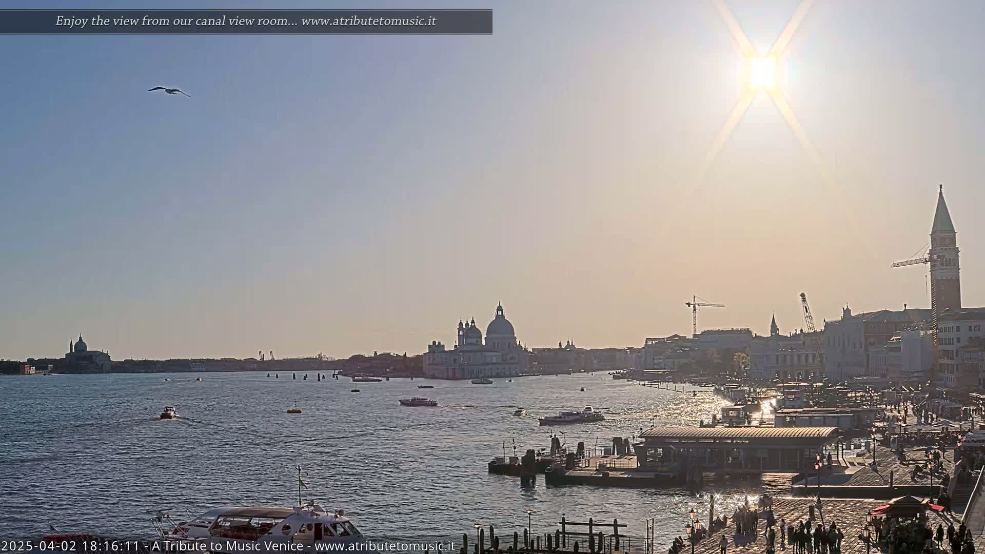 A sunny view across a Venetian canal shows boats on the water, a cityscape with several domes and a tall bell tower, and people walking along a paved waterfront.