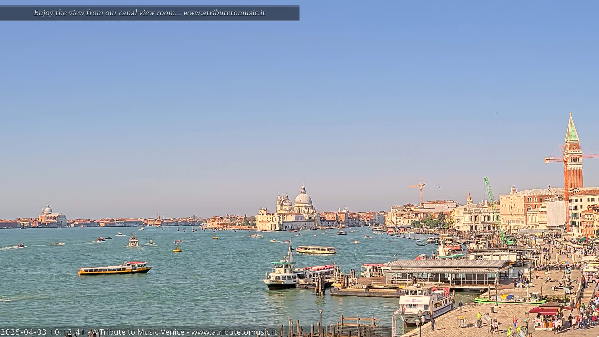 A sunny view of a Venetian canal shows several boats, a dock, and buildings including a large domed church, and a tall red brick campanile.