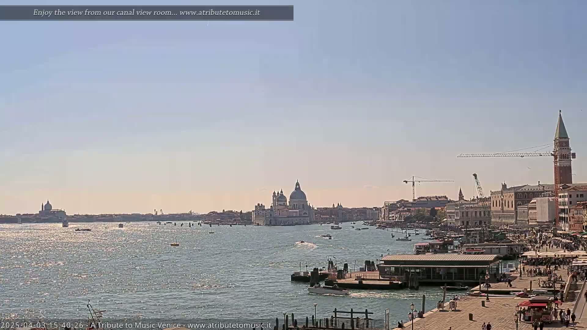 A sunny day in Venice shows a wide canal with boats, buildings, and a large church in the distance, under a clear sky.