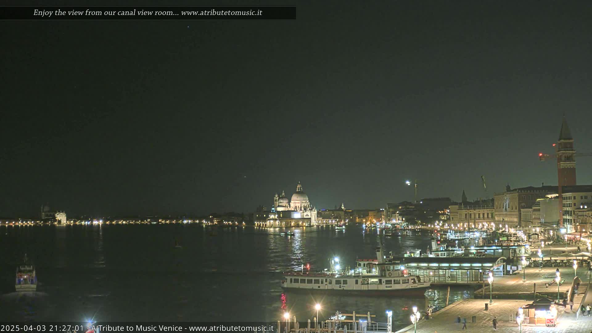 A nighttime view of a canal in Venice, showing illuminated buildings and boats, under a dark sky.