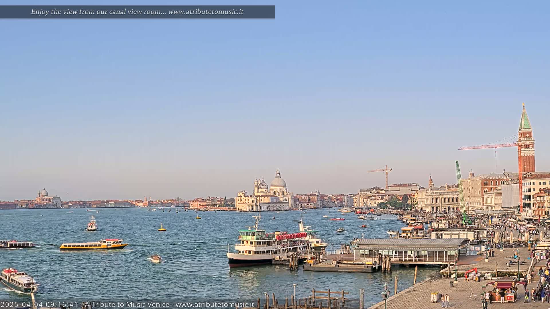 A sunny day in Venice shows a wide canal with numerous water taxis and passenger ferries, a city skyline featuring a large domed church, and a bustling waterfront area with people and buildings.