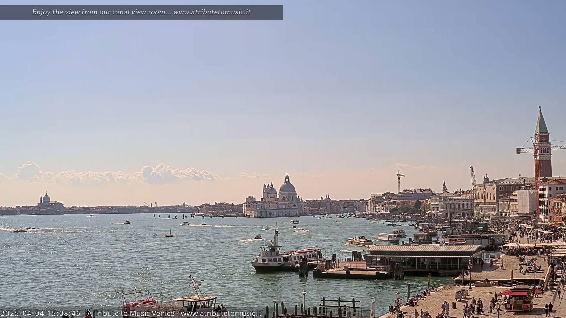A sunny day in Venice shows a canal with boats, a waterfront promenade filled with people, and the Basilica di Santa Maria della Salute in the distance.