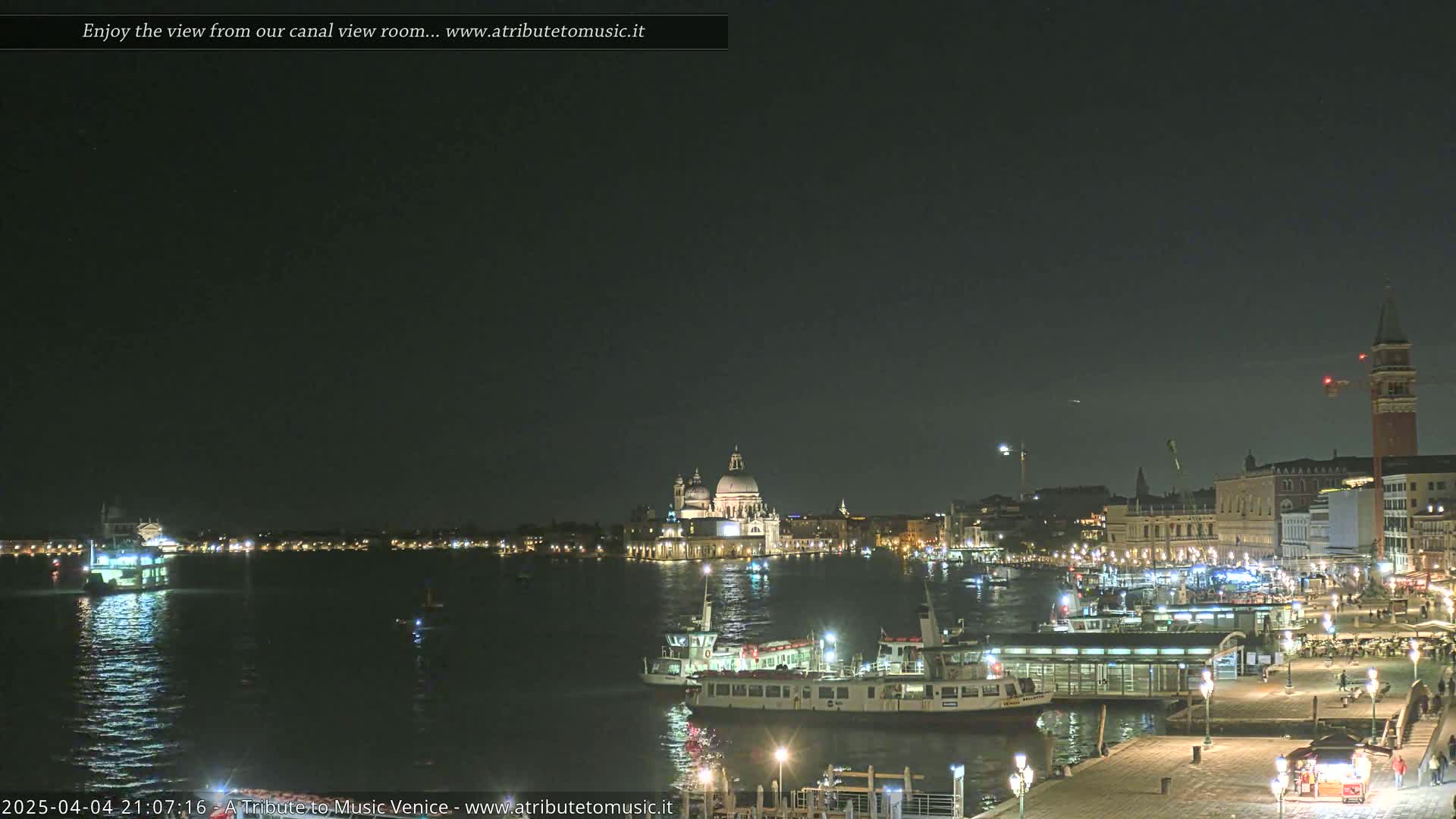 A nighttime view across a calm canal shows illuminated buildings and boats in Venice, under a dark sky.