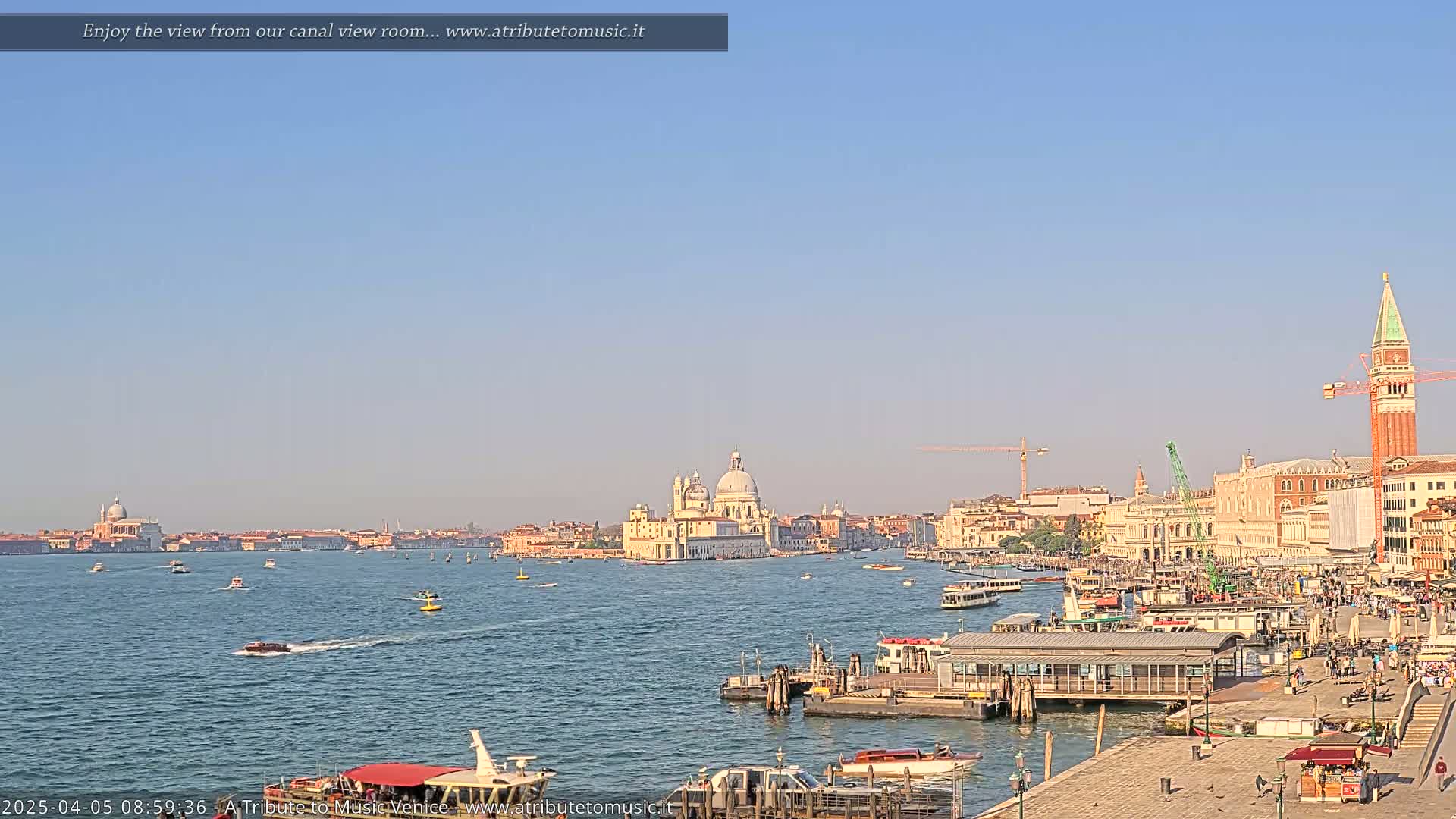 A clear, sunny day reveals a wide canal in Venice, Italy, with numerous boats and buildings lining the waterway, including a large church-like structure and a tall bell tower.