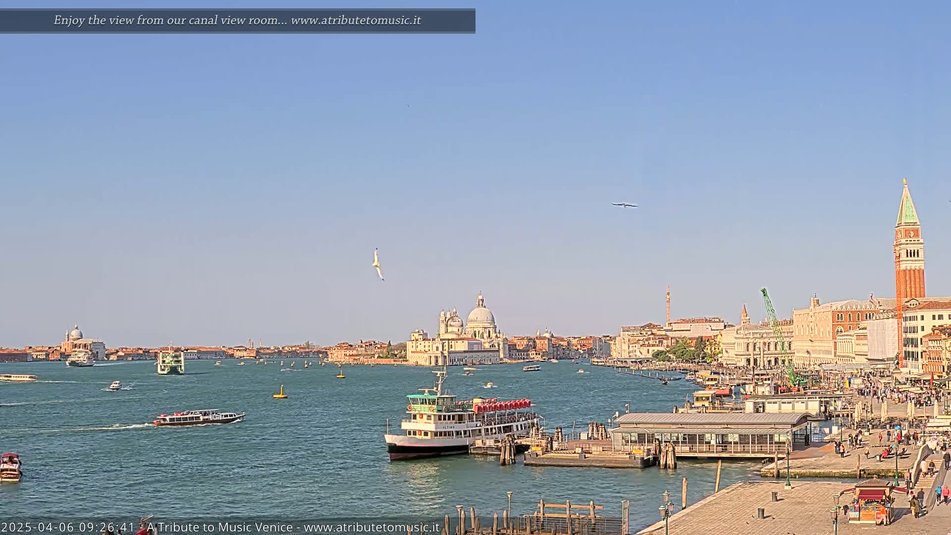 A sunny day in Venice shows a wide canal with several boats, a large passenger ferry at a dock, and the city's buildings and landmarks in the background.