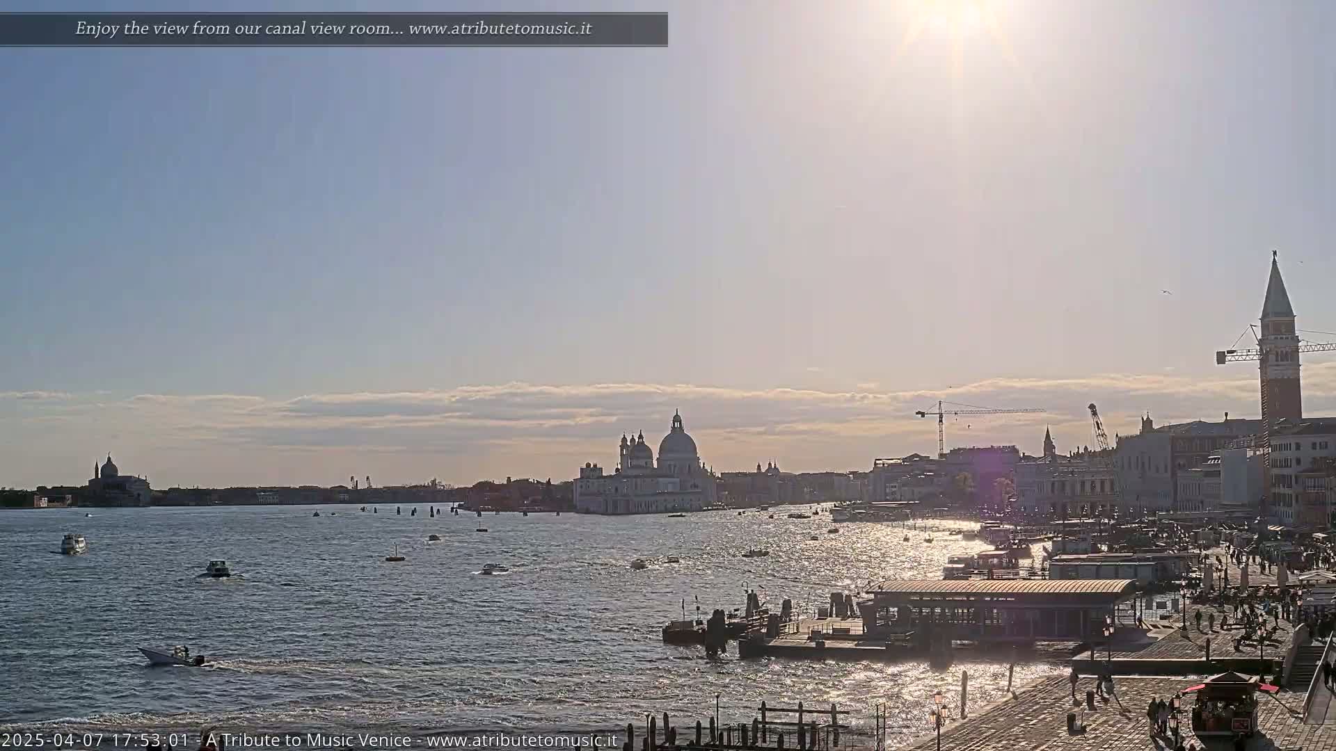 A sunny, partly cloudy day in Venice shows a wide canal with boats, and a cityscape including a large domed church and a tall bell tower.