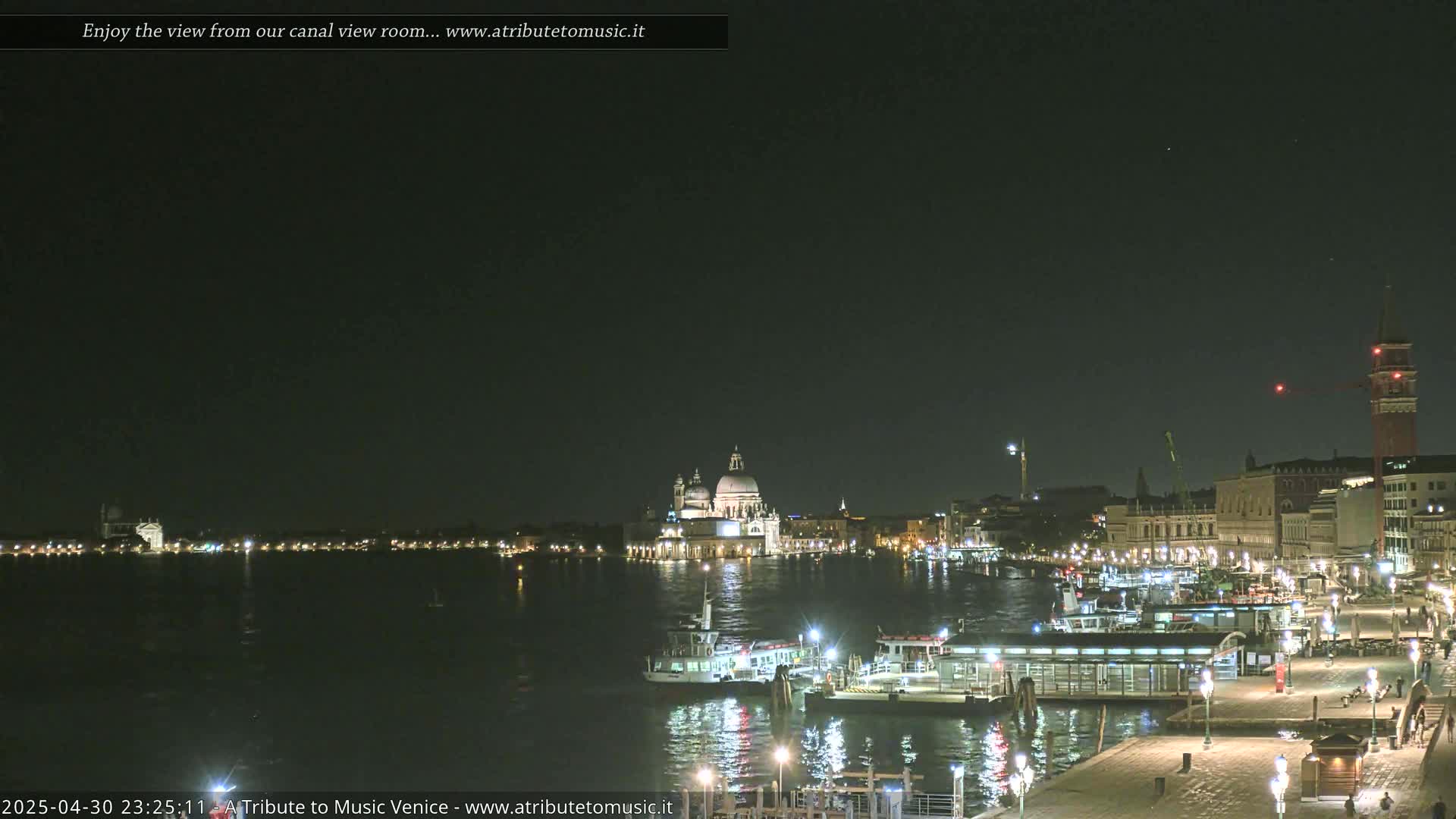 A nighttime view of a canal in Venice, Italy, shows illuminated buildings and boats on calm water under a dark sky.