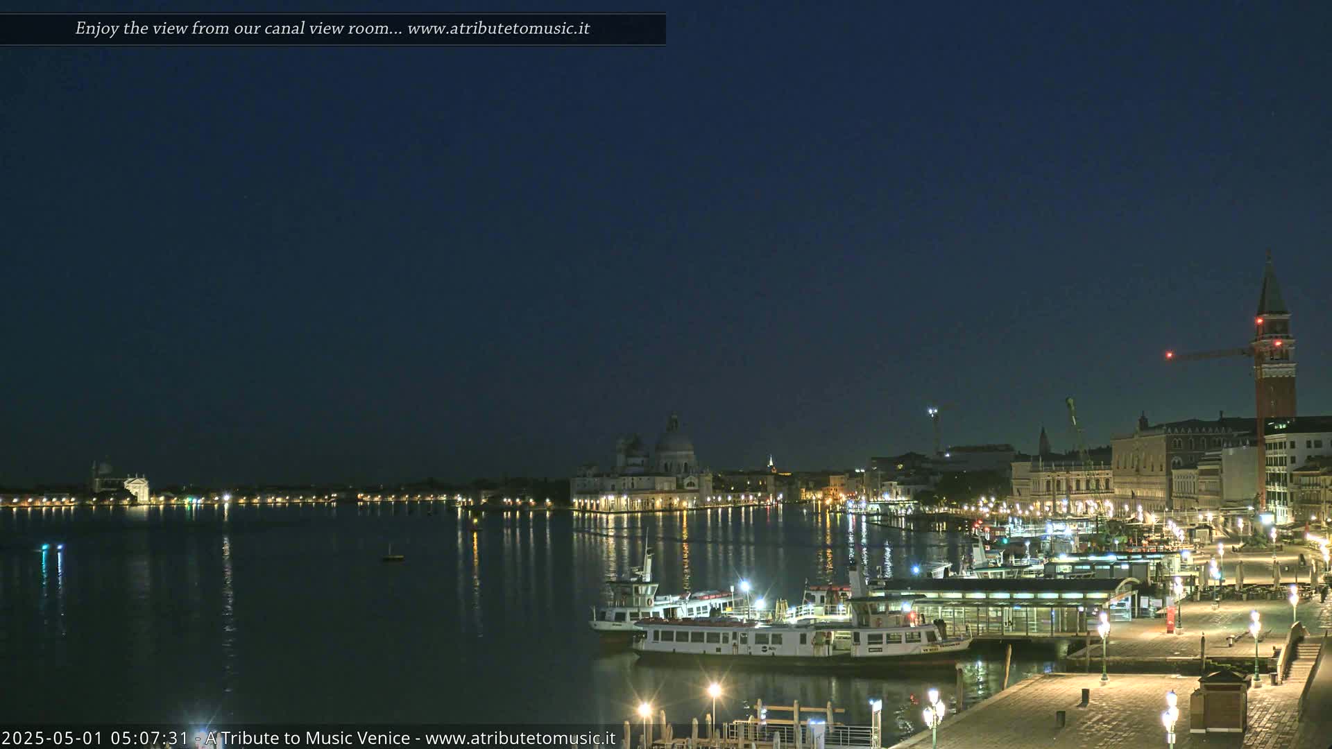 A nighttime view across a calm body of water shows illuminated buildings, boats, and a pier under a dark, clear sky.