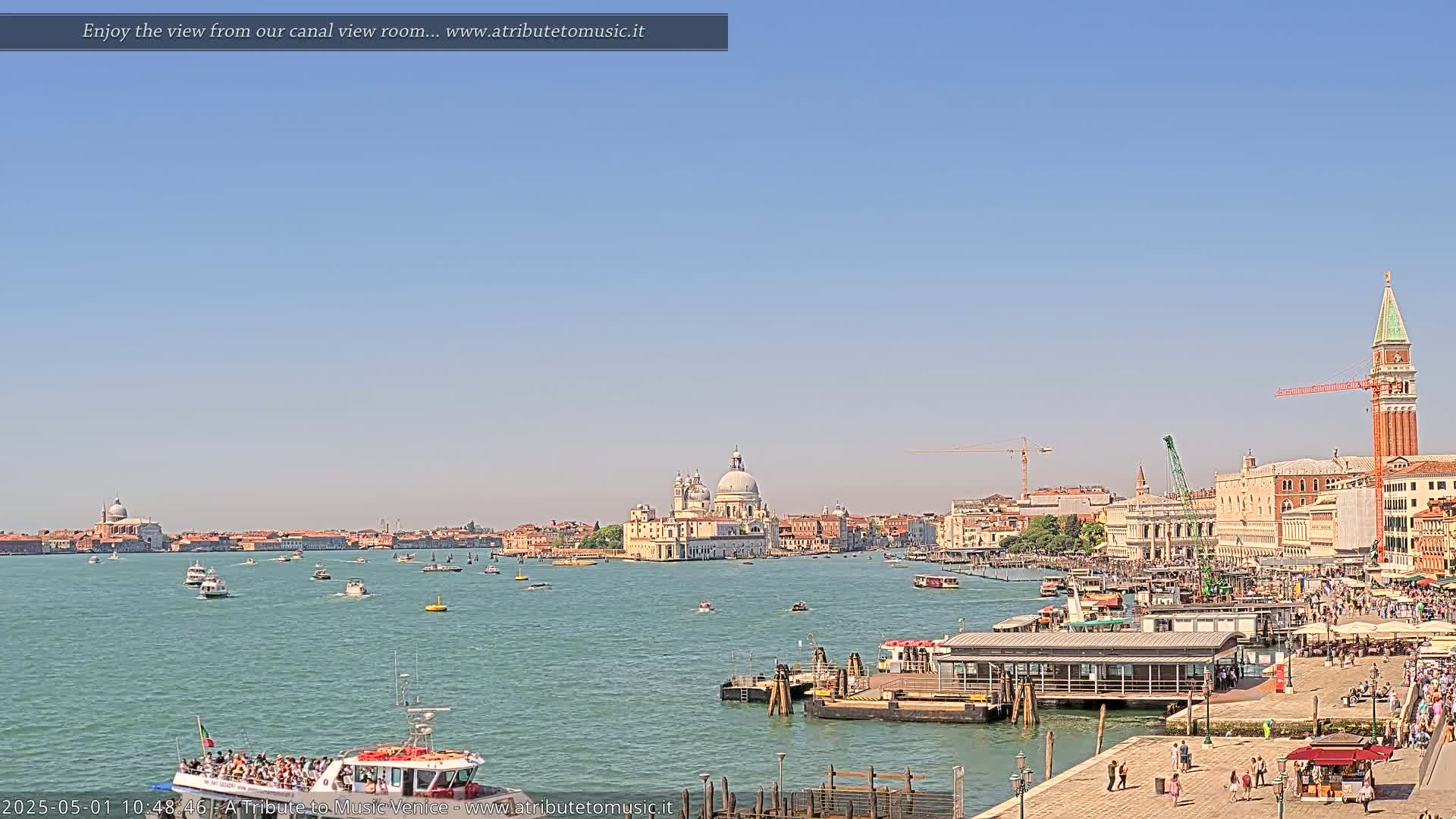 A sunny view of Venice, Italy, shows a wide canal with numerous boats, a large church in the distance, and a waterfront area with people and buildings.