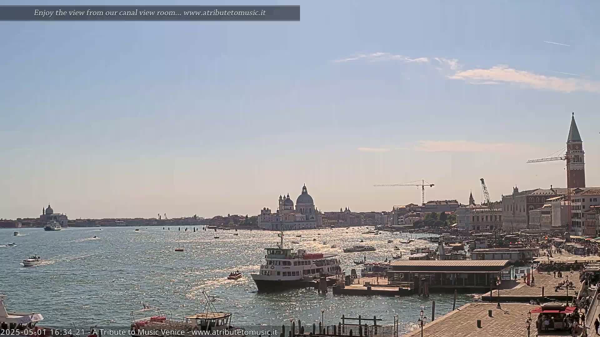 A sunny day in Venice shows a wide canal with boats, a large church in the distance, and a waterfront area with buildings and people.