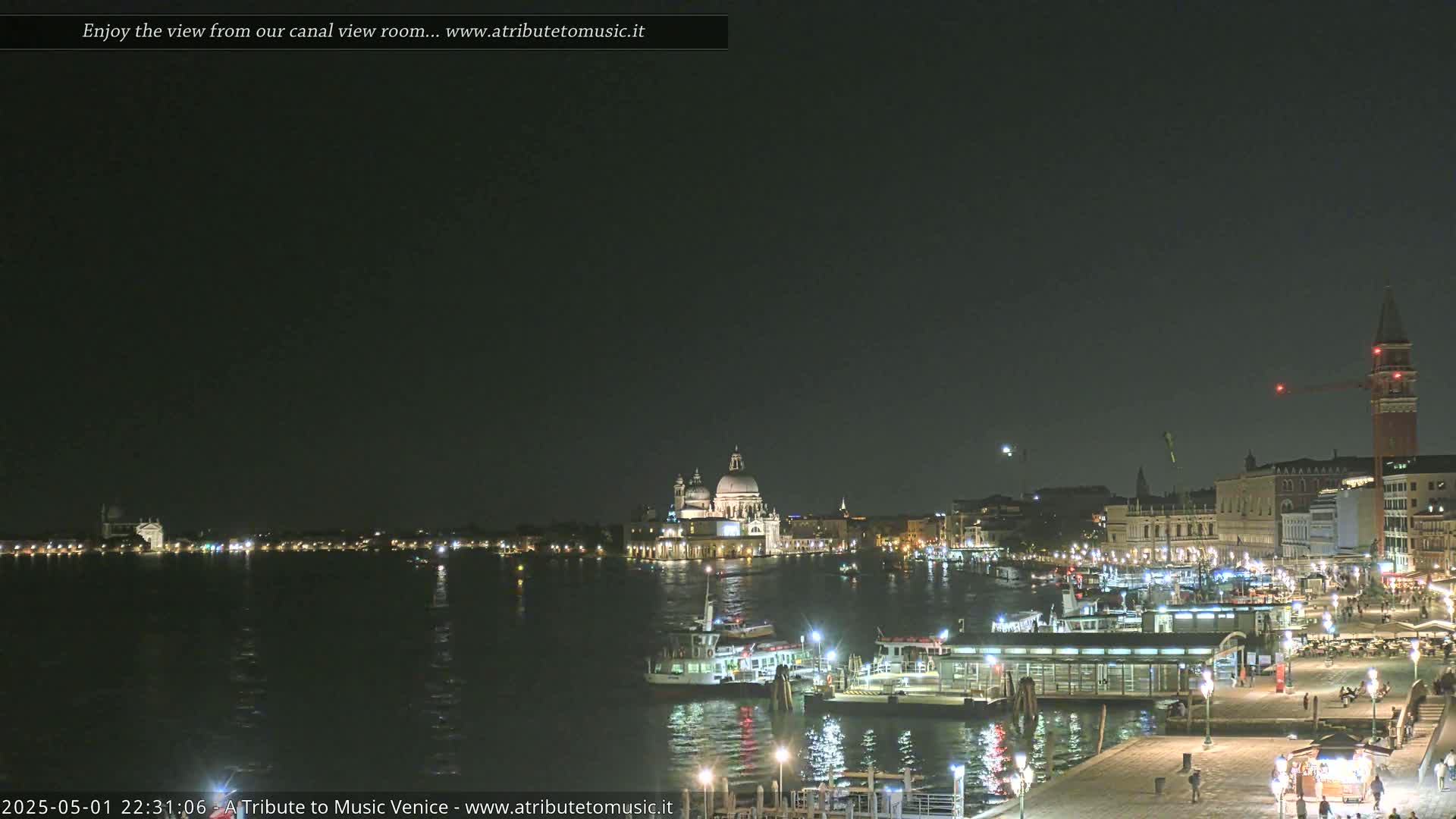 A nighttime view of a canal in Venice, Italy, shows illuminated buildings and boats, with a clear, dark sky.