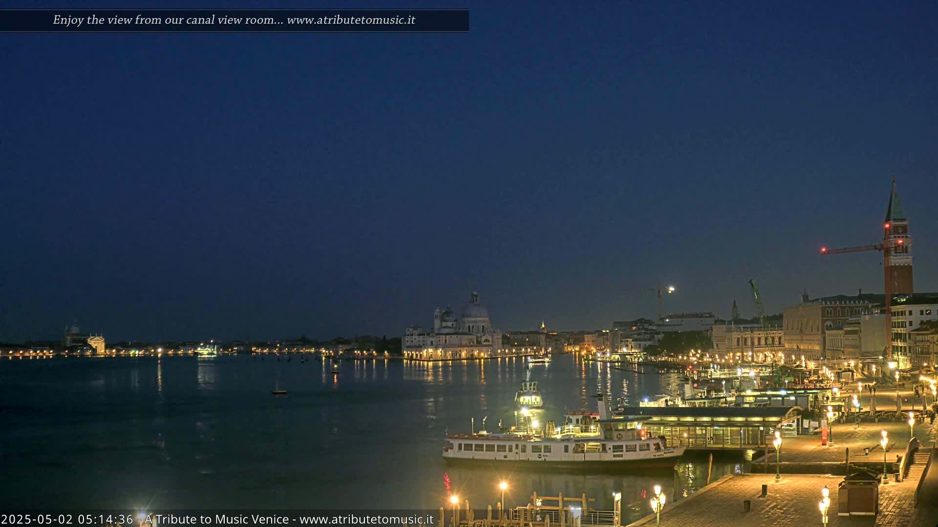 A nighttime view across a calm canal shows illuminated buildings and a ferry, under a dark blue sky.