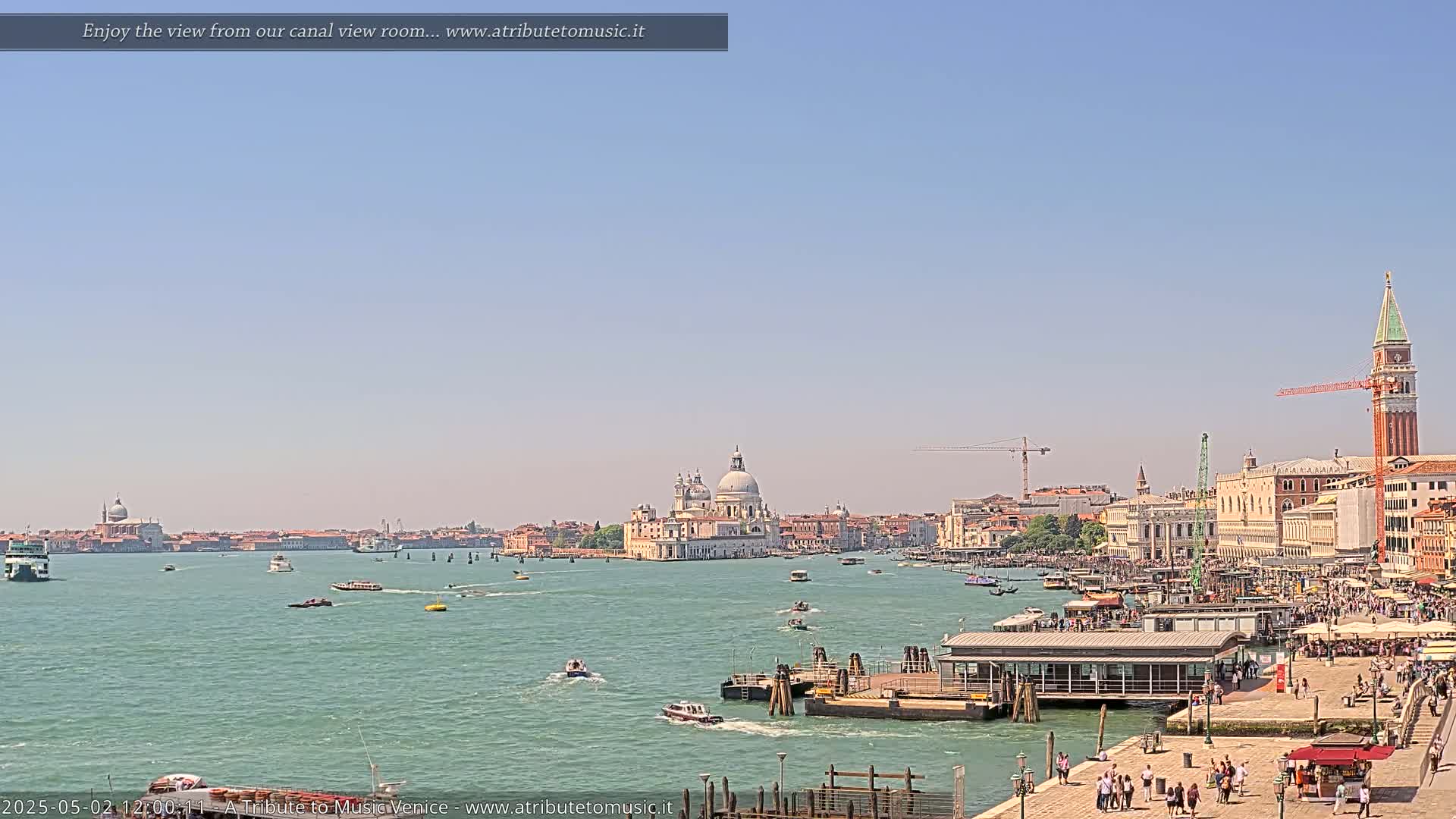 A sunny day in Venice shows a wide canal with boats, a waterfront promenade filled with people, and the Basilica di Santa Maria della Salute in the distance.