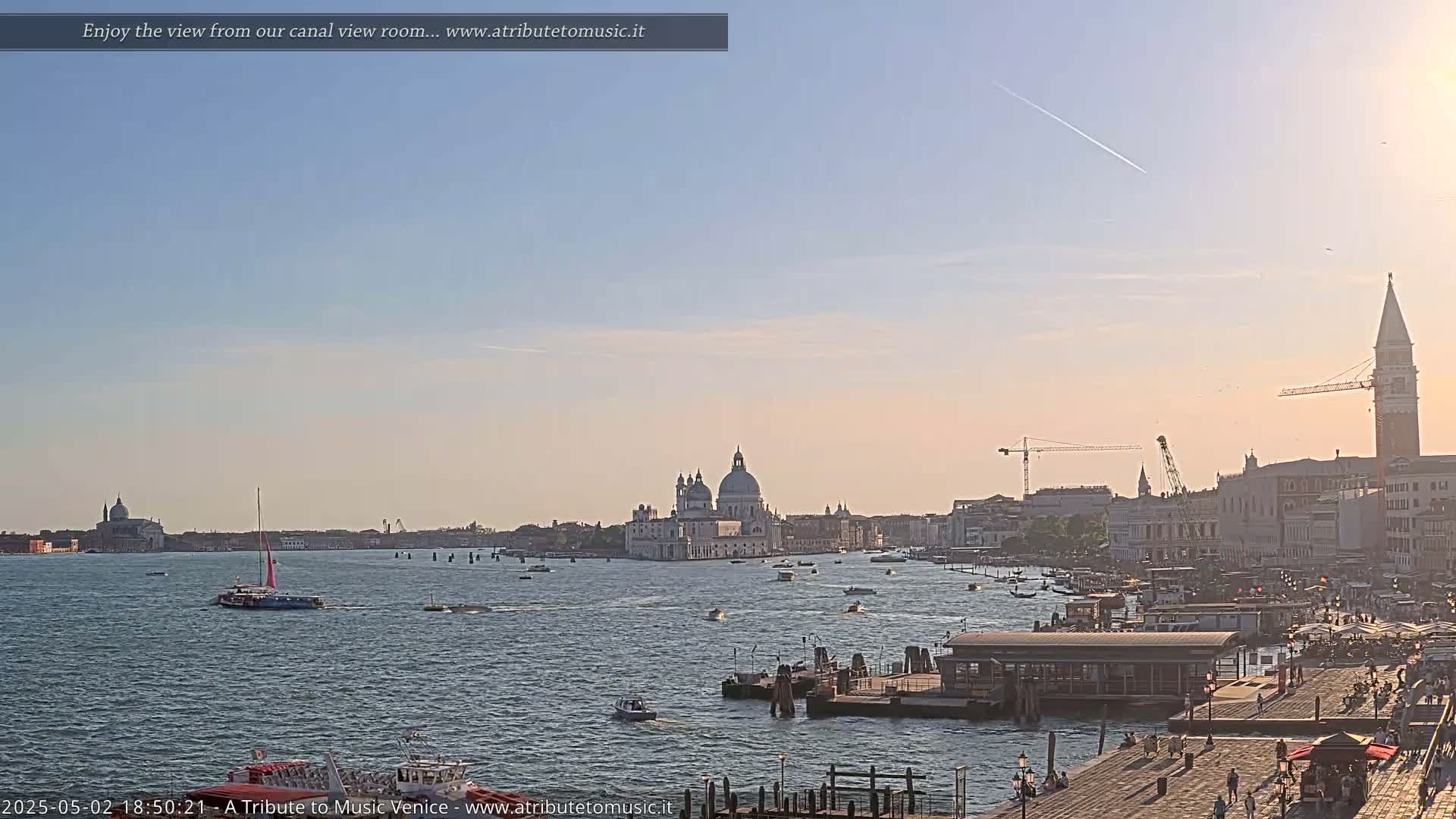 A wide shot of a Venetian canal at sunset shows numerous boats on the water, buildings lining the canal, and a clear sky with a faint contrail.