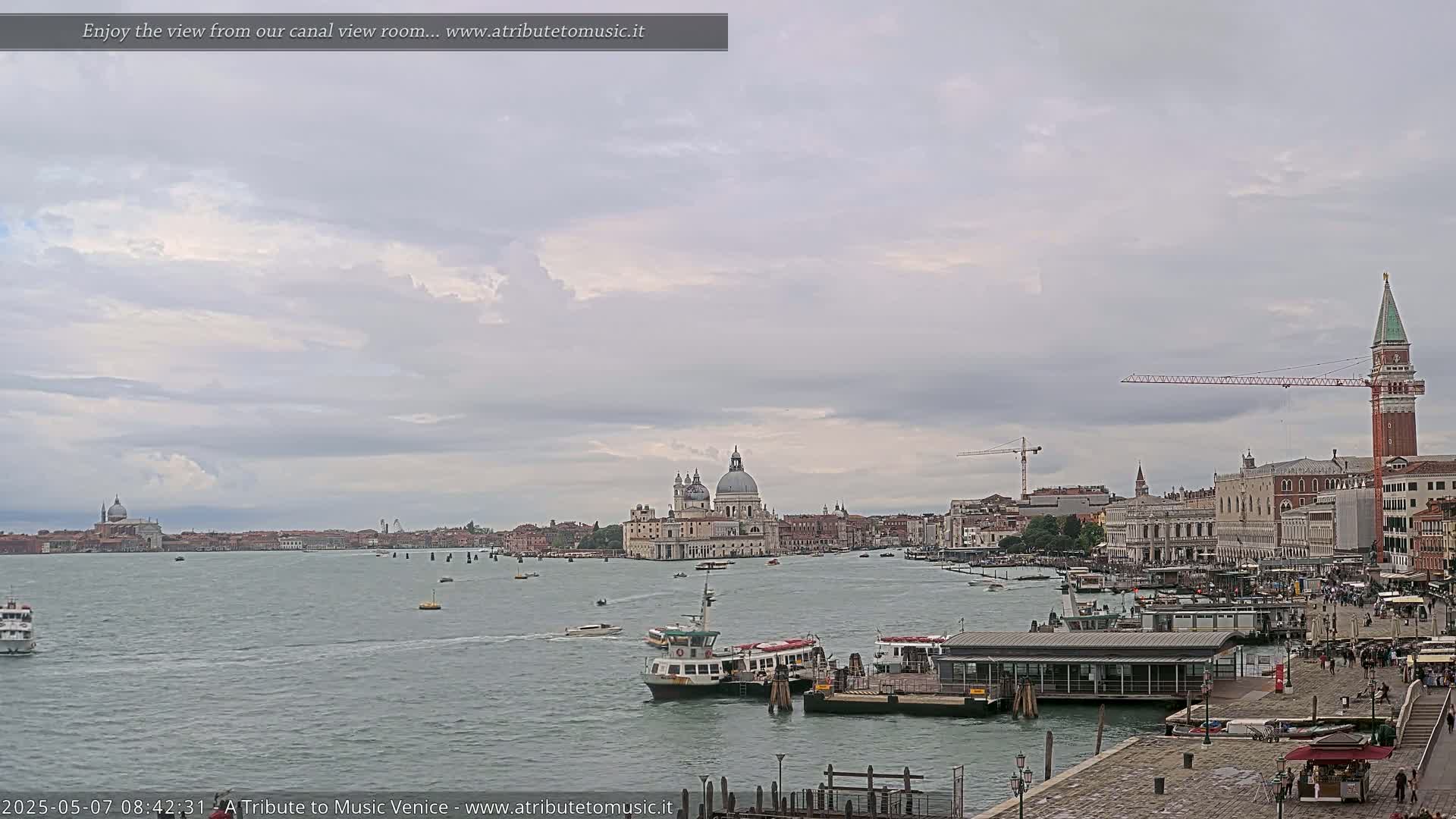A wide view of a waterway in Venice, under a cloudy sky, shows several boats and buildings including a large church, with a crane visible in the distance.