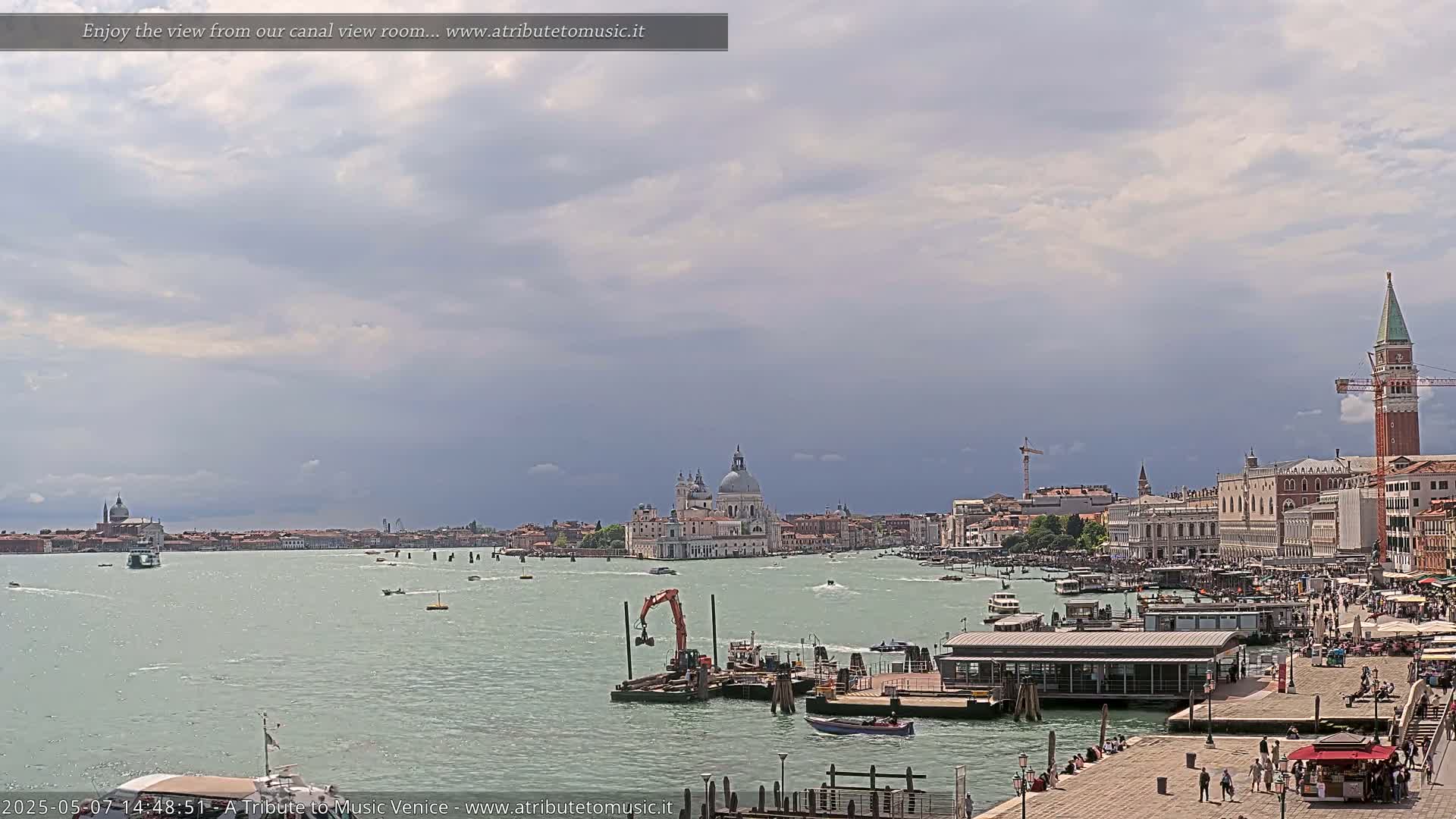 A wide shot of a Venetian waterway shows several boats, a construction barge, buildings, and a large church under a partly cloudy sky.
