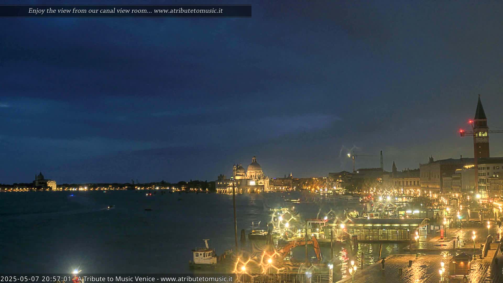 A nighttime view of a canal in Venice, showing illuminated buildings and boats under a dark blue sky.