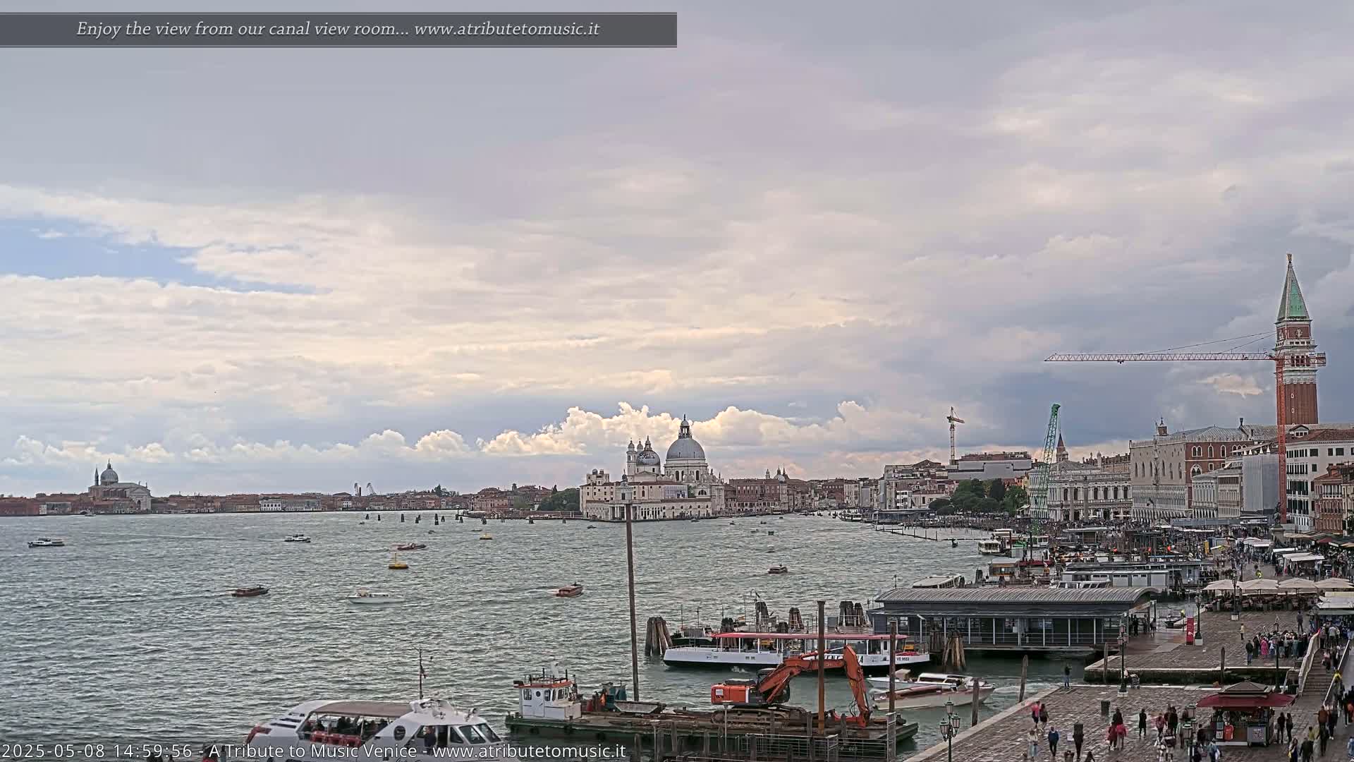 A wide shot of a Venetian waterway shows boats, a city skyline featuring a large domed basilica, a tall bell tower, and construction cranes under a partly cloudy sky.