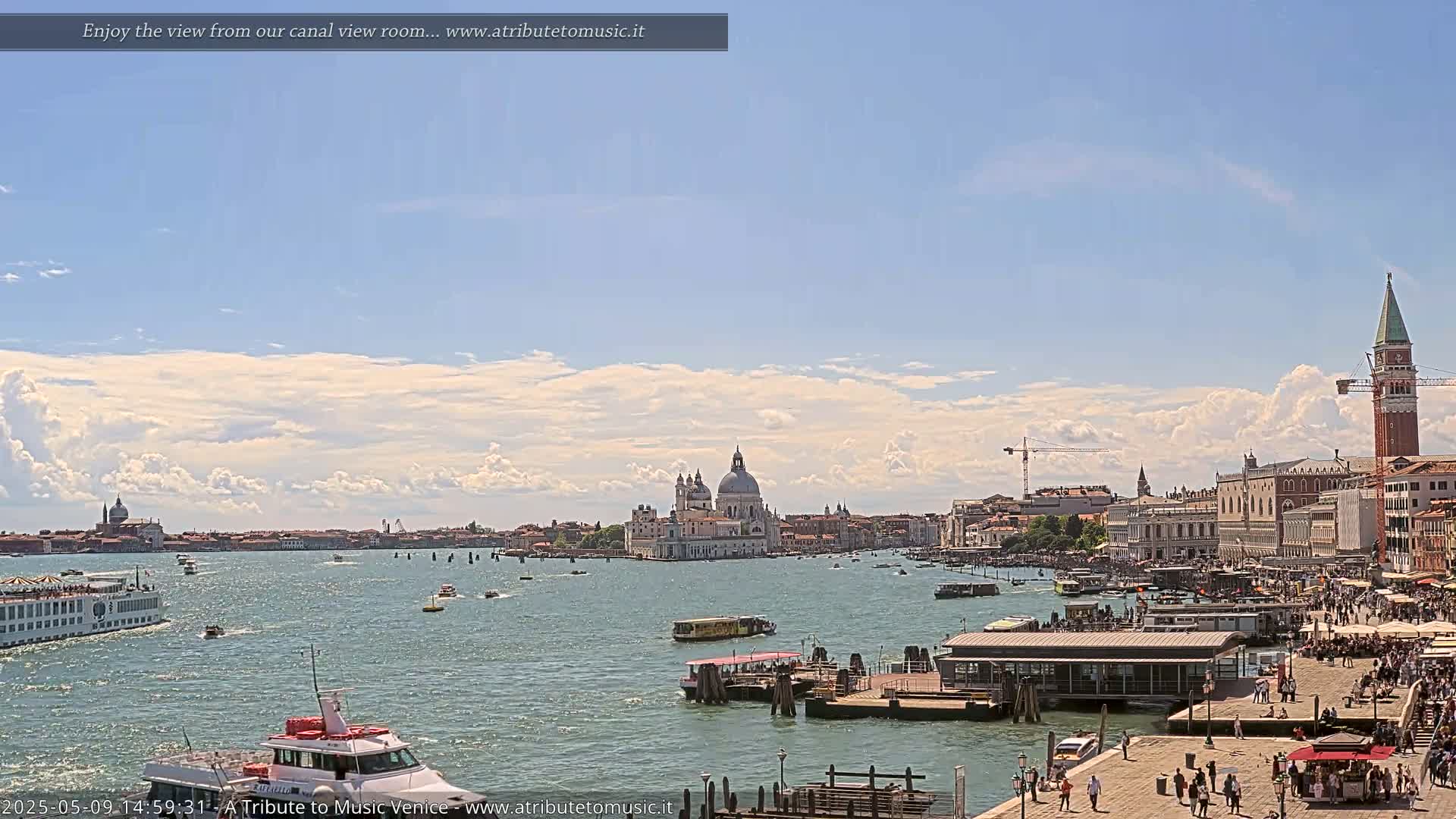 A sunny day in Venice shows a wide canal with boats, a large building on an island in the middle, and a crowded waterfront with buildings and a bell tower.