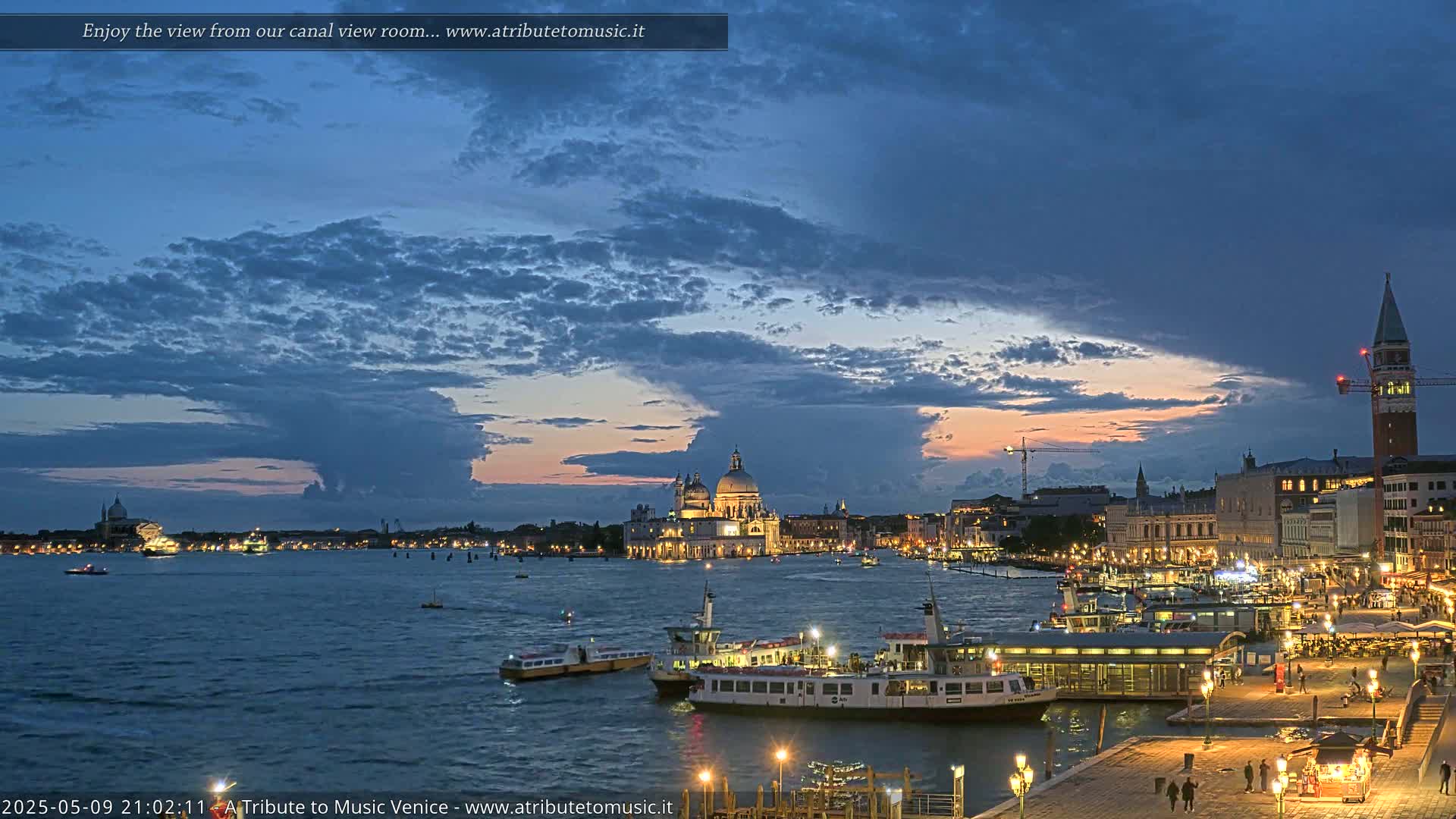 A nighttime view of a Venetian canal with boats, illuminated buildings, and a cloudy twilight sky.