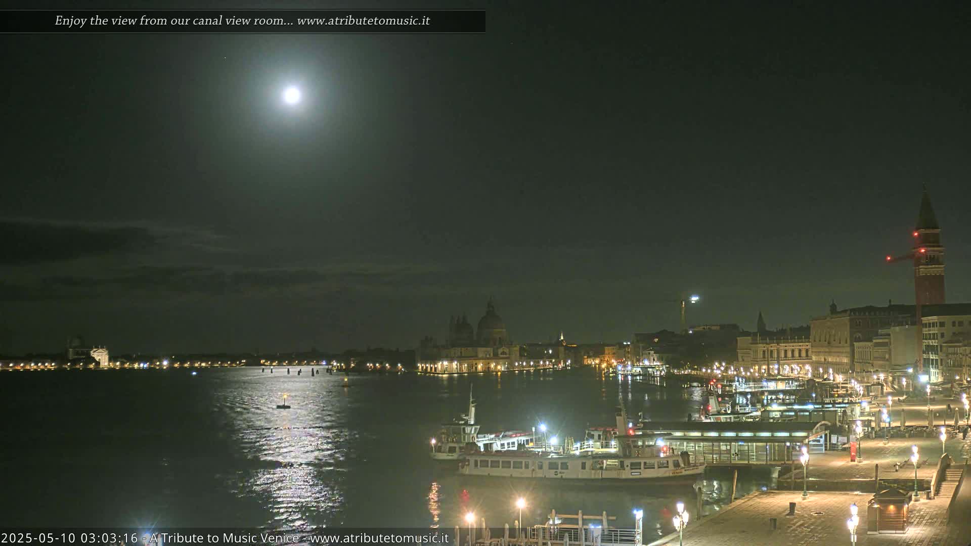 A nighttime view of a moonlit canal in Venice, showing illuminated buildings, boats, and a pier.