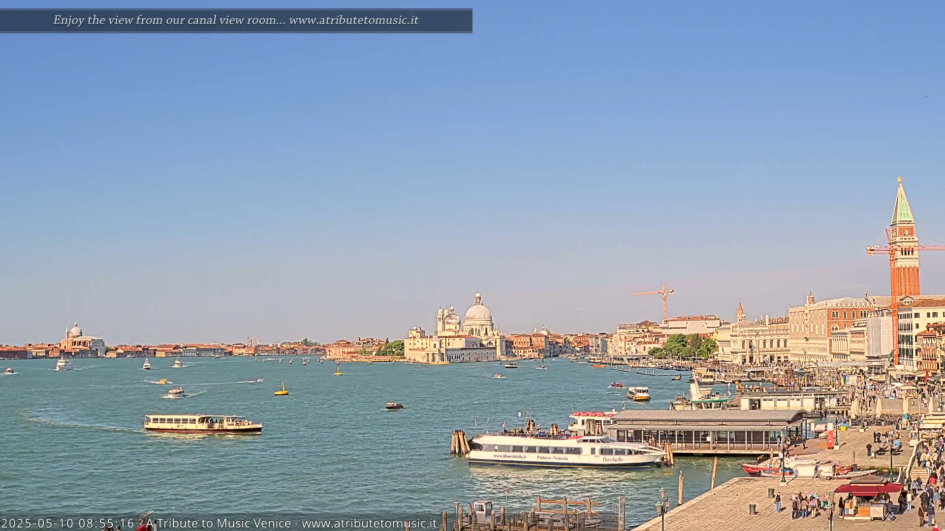 A sunny day in Venice shows a wide canal with several boats, buildings lining the waterfront, and the San Giorgio Maggiore church visible in the distance.
