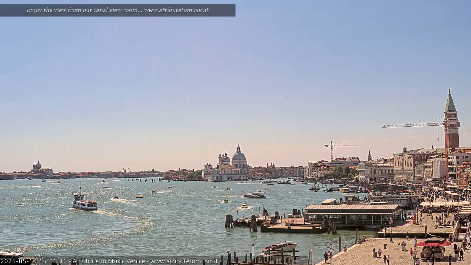 A sunny view across a calm waterway shows a Venetian cityscape with boats, a large church, and a tall bell tower under a clear blue sky.